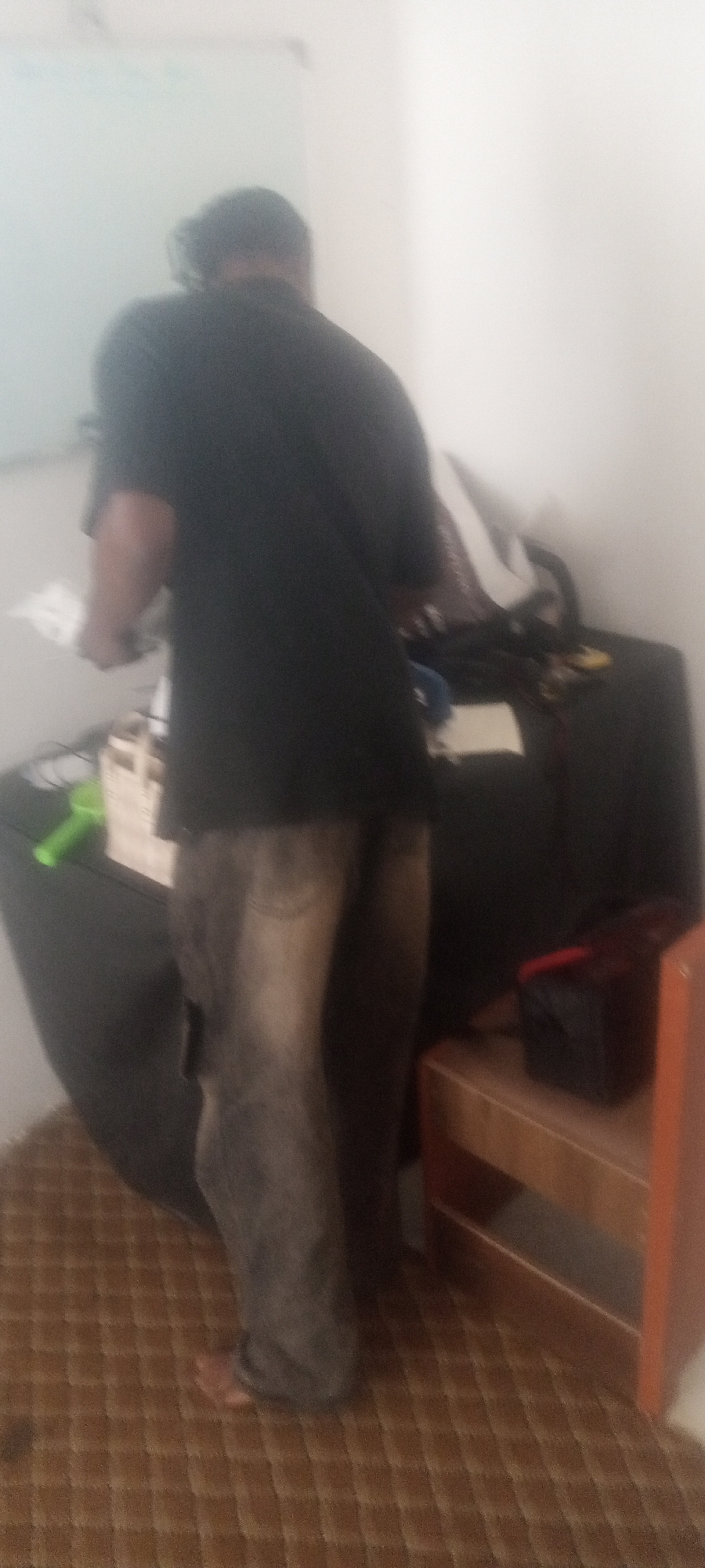 A blurry, indoor photograph shows a person standing near a table and a whiteboard. The person, seen from the back, wears a dark t-shirt and faded jeans. They appear to be holding something in their hands. Behind the person is a dark table or cabinet, cluttered with various objects. To the right of the table is a small, wooden shelf. The floor has a textured, possibly carpeted, surface with a grid-like pattern. The lighting is dim, and the image is generally out of focus.