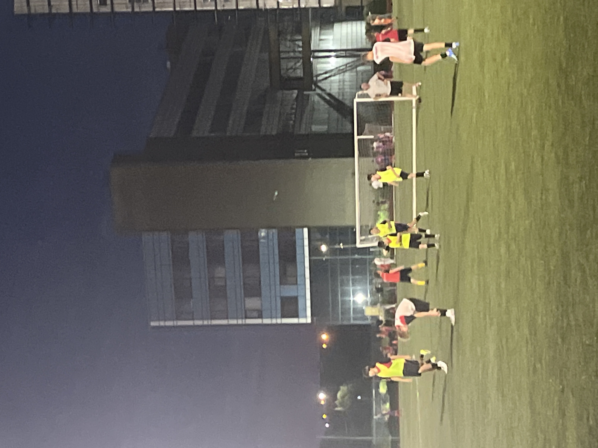 In this vertically oriented image, a group of people are playing soccer on a grass field at night. The scene is illuminated by artificial lights, and the sky is dark, suggesting it is nighttime. Behind the soccer field, there are modern buildings with multiple stories. One building is a tall, dark structure with many windows, and another is shorter with blue and white panels. A soccer goal is visible on the field.
The individuals are wearing athletic clothing. Some are wearing yellow vests over their shirts, and others are in red or pink shirts. Their postures and positions suggest they are actively engaged in a soccer game or practice, with some appearing to be running or in motion. One person in a pink shirt is bending over. The overall atmosphere appears to be one of athletic activity. There are no visible signs or text in the image. The setting appears to be an urban area in Krasnodar, Russia, given the presence of the modern buildings and the grassy field.