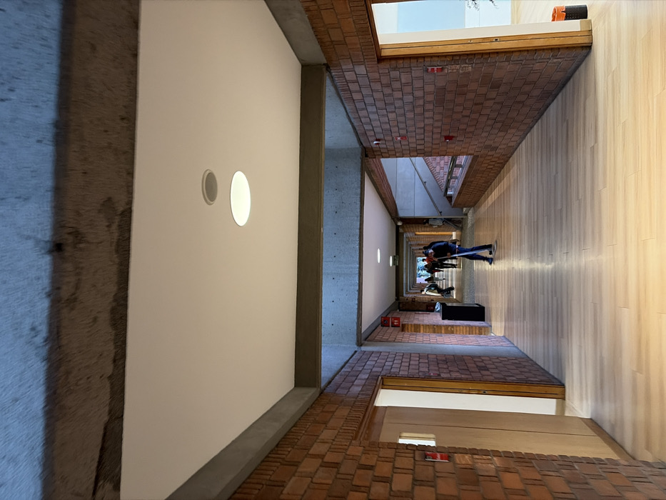 The image displays an interior view of a long, illuminated institutional corridor located within a building at the National Autonomous University of Mexico (UNAM) in Ciudad Universitaria, Coyoacán, Mexico City. The corridor features a light-toned wooden floor, reddish-brown brick walls arranged in a stretcher bond pattern, and a white dropped ceiling equipped with circular recessed light fixtures and a smaller recessed speaker. Above the dropped ceiling and along its perimeter, rough, exposed concrete beams and a concrete ceiling are visible.
In the mid-foreground, slightly right of center, a person dressed in dark clothing is actively sweeping or mopping the wooden floor with a long-handled implement. Further down the corridor, several other individuals are visible walking away from the camera, their features obscured by distance.
On the left wall, a light-colored wooden door is present in the foreground, followed by a black waste receptacle and a pair of red restroom pictograms (male and female symbols). Red wall-mounted devices, consistent with fire alarms or emergency buttons, are spaced along both brick walls. On the right side of the corridor, an open doorway reveals a brightly lit internal space. Further down the right, a brick half-wall topped with concrete runs alongside a visible staircase, indicating an adjacent multi-level area. The overall environment suggests an active academic or administrative setting.