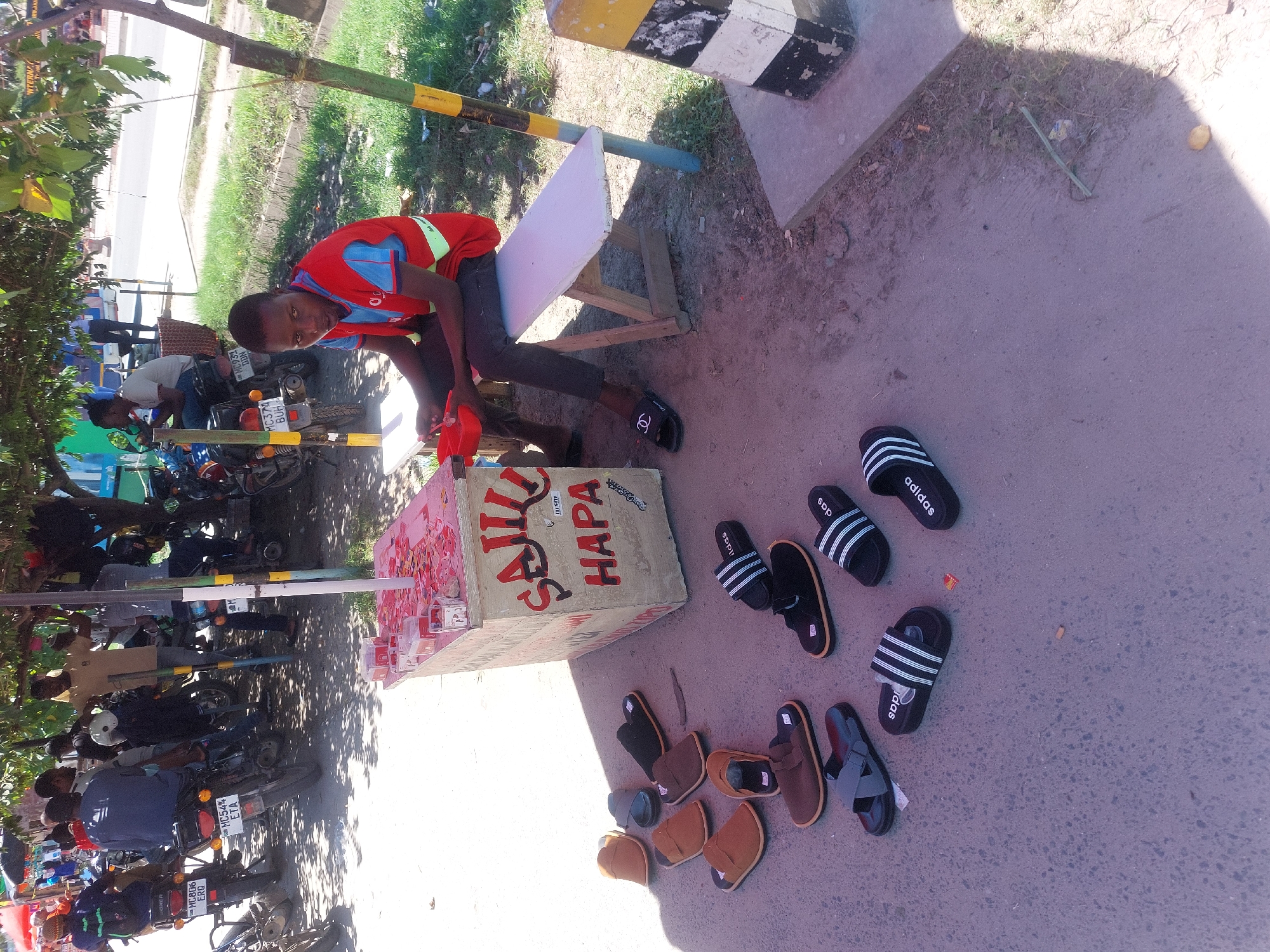 A youth sits on a bench at an informal roadside stall in Dar es-Salaam, Tanzania, near a curb. The stall displays sandals, with Adidas and similar styles scattered on the ground. The stall is painted with the words "SAJU HAPA." Motorcycles are parked along the street under a tree. The youth is wearing a red shirt and dark pants with Chanel-branded sandals. A striped yellow and black pole stands adjacent to the bench.