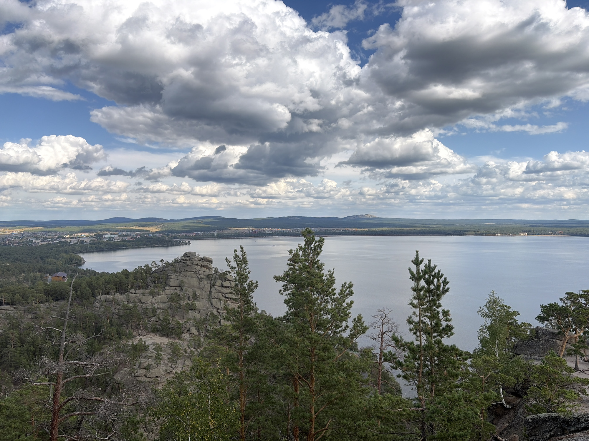 The image captures a scenic overview of the city of Zerenda, located in the Zelionoborskiy rural district of Kazakhstan. In the foreground, a rocky outcrop is densely covered with pine trees, some appearing dry and skeletal. A larger, weathered rock formation dominates the mid-ground, with trees clinging to its slopes. Beyond this, a tranquil lake stretches out, its surface reflecting the dramatic, cloud-filled sky. On the far shore of the lake, the city of Zerenda is visible, with buildings clustered along the waterfront and nestled amongst the trees. The background features rolling hills and a distant mountain range under a vast expanse of sky characterized by a mix of bright white and dark gray clouds. The overall scene conveys a sense of natural beauty and the integration of human settlement within a rugged landscape.