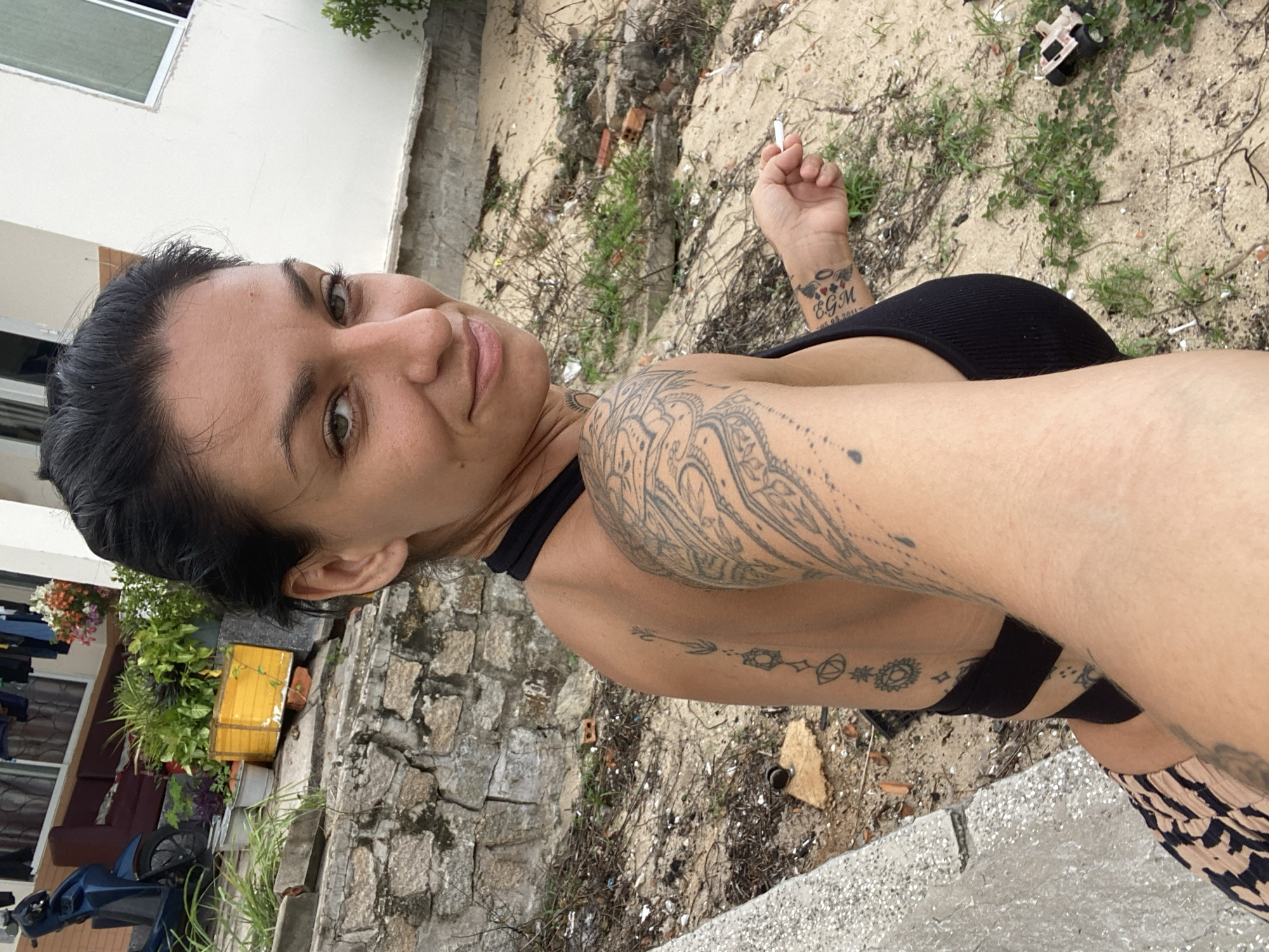 A woman is captured in a selfie, her body angled to the left and her head turned to face the camera. She has dark hair pulled back, and her gaze is directed towards the viewer with a slight smile. She is wearing a black halter top and a patterned skirt. Tattoos adorn her left arm and back. In her right hand, she holds a cigarette. The background appears to be a sandy, somewhat overgrown area with scattered debris, including bricks and a small toy vehicle. A stone wall is visible to the left, and a white building stands in the background. The scene suggests an outdoor setting in Mũi Né, Vietnam, possibly a less developed area.