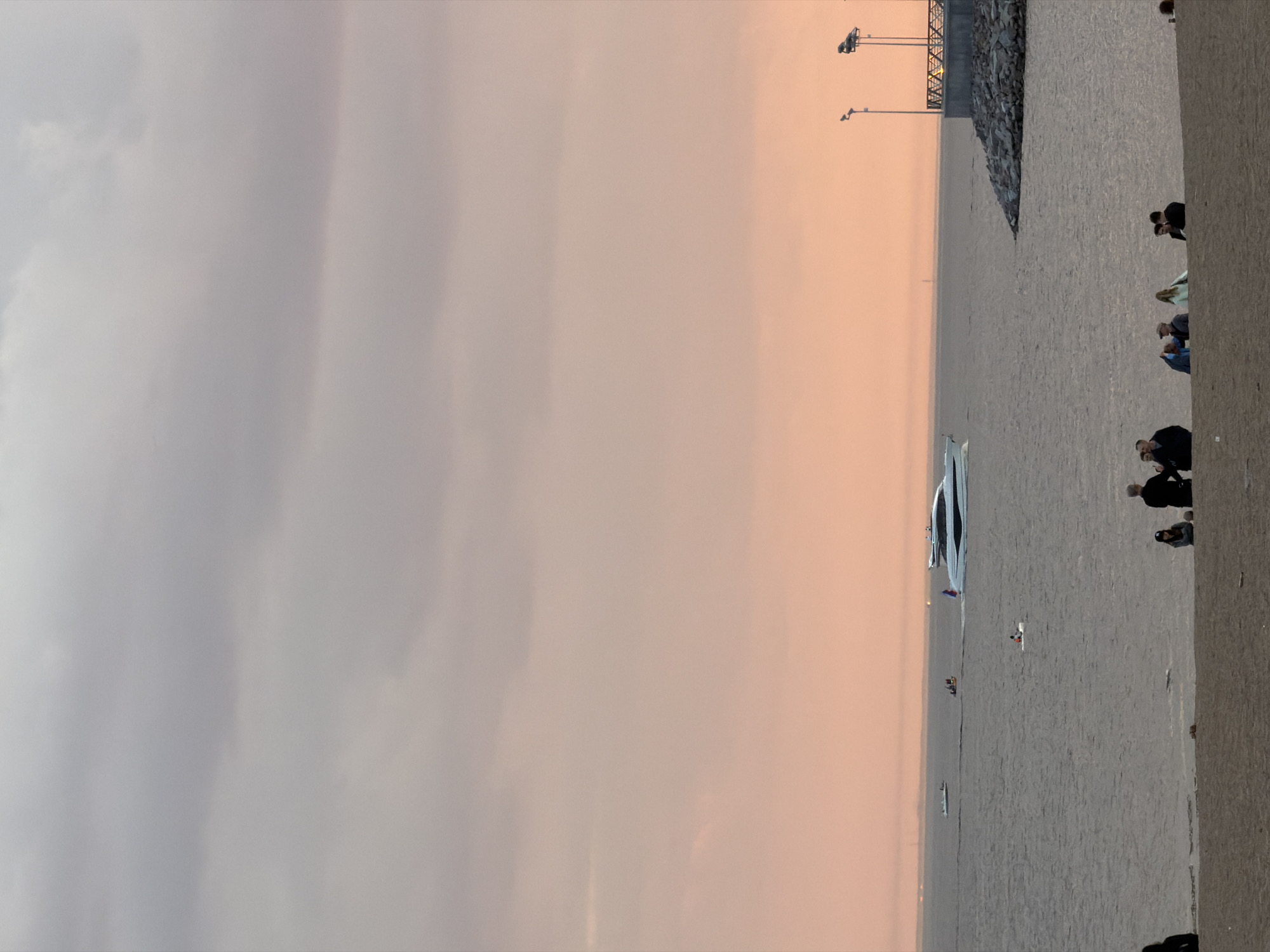 This media file captures a serene waterfront scene, likely a promenade or pier, at either dawn or dusk. The sky presents a soft gradient of colors, transitioning from a light gray-blue on the left to a warm, gentle orange-pink towards the right, indicative of a rising or setting sun. Overcast or diffuse cloud cover is visible, especially on the left side of the sky.
On the right side of the image, a wide, paved walkway or promenade extends into the distance. Several small groups of people are scattered along this area; they appear to be casually dressed, standing or sitting, and observing the water. Their specific features are not discernible due to the distance.
The expansive body of water, likely a bay or gulf near Saint Petersburg, Russia, shows subtle ripples. A prominent, sleek white yacht or large boat is visible in the mid-ground on the water. Further out, a lone individual in a small white kayak or similar vessel is seen, and a few other distant boats or structures can be faintly made out on the horizon. To the far right, a rocky embankment or breakwater is present, behind which industrial-looking structures or tall light poles are silhouetted against the sky. The overall atmosphere is tranquil and relaxed. No visible text is present in the image.