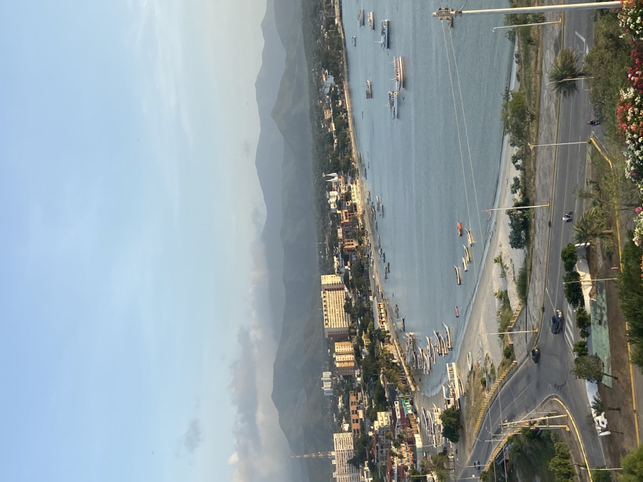 A high-angle shot captures a coastal scene. Mountains rise in the background, meeting a shoreline dotted with buildings. Boats populate the calm water alongside a beach, while a road runs parallel to the coast. Cars move along the road, which is bordered by greenery and trees. Utility poles and power lines are visible. The sky is clear and light blue.