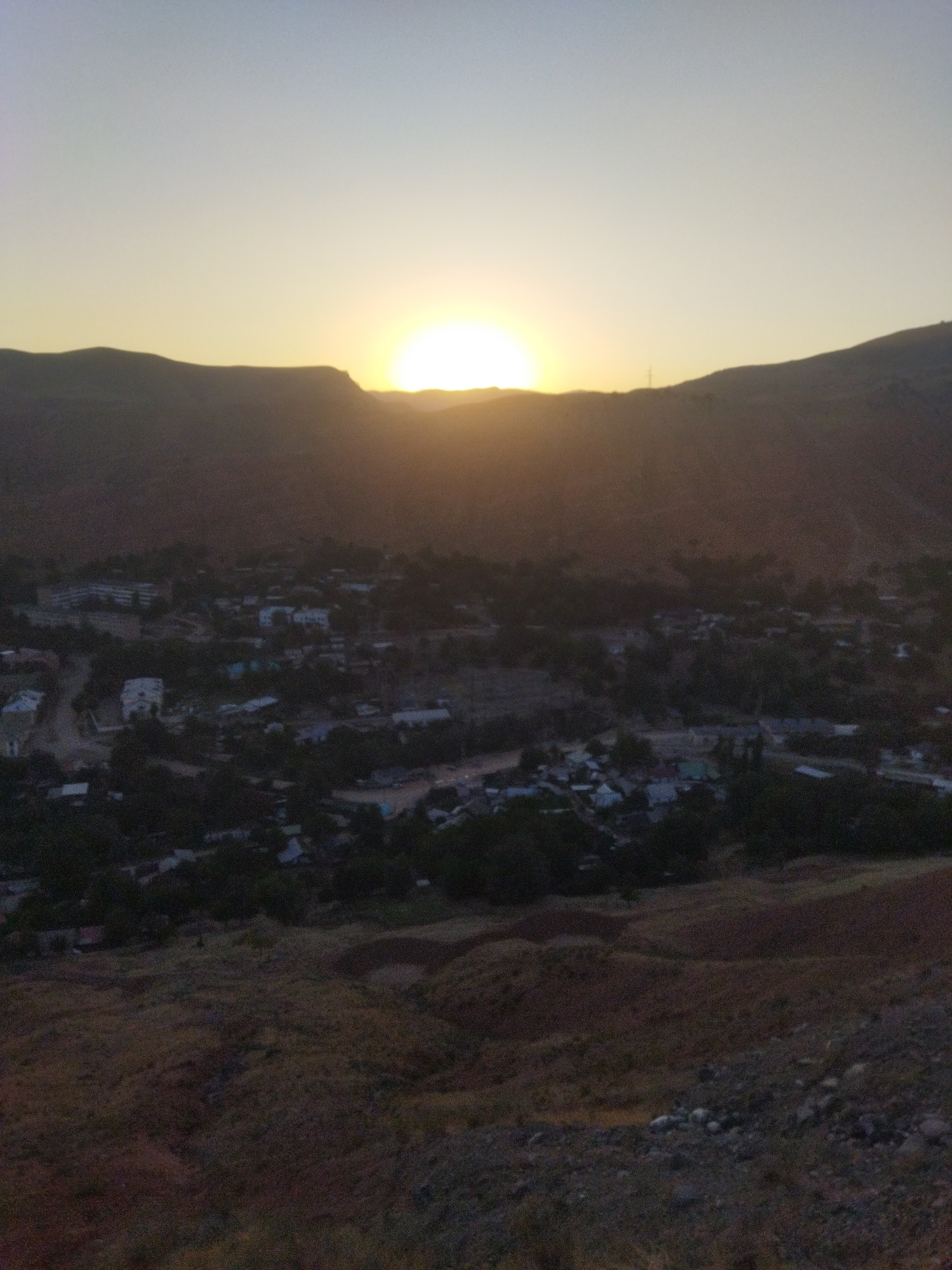 This image captures a panoramic view of Mailuu-Suu City, Kyrgyzstan, during a sunset. The sun is partially obscured by a mountain range in the background, casting a warm, golden glow across the sky. In the midground, a cluster of buildings, indicative of a residential area, is nestled amongst trees. The foreground shows a rugged, hilly terrain with sparse vegetation. The overall atmosphere is serene, with the setting sun creating long shadows and a diffused light.