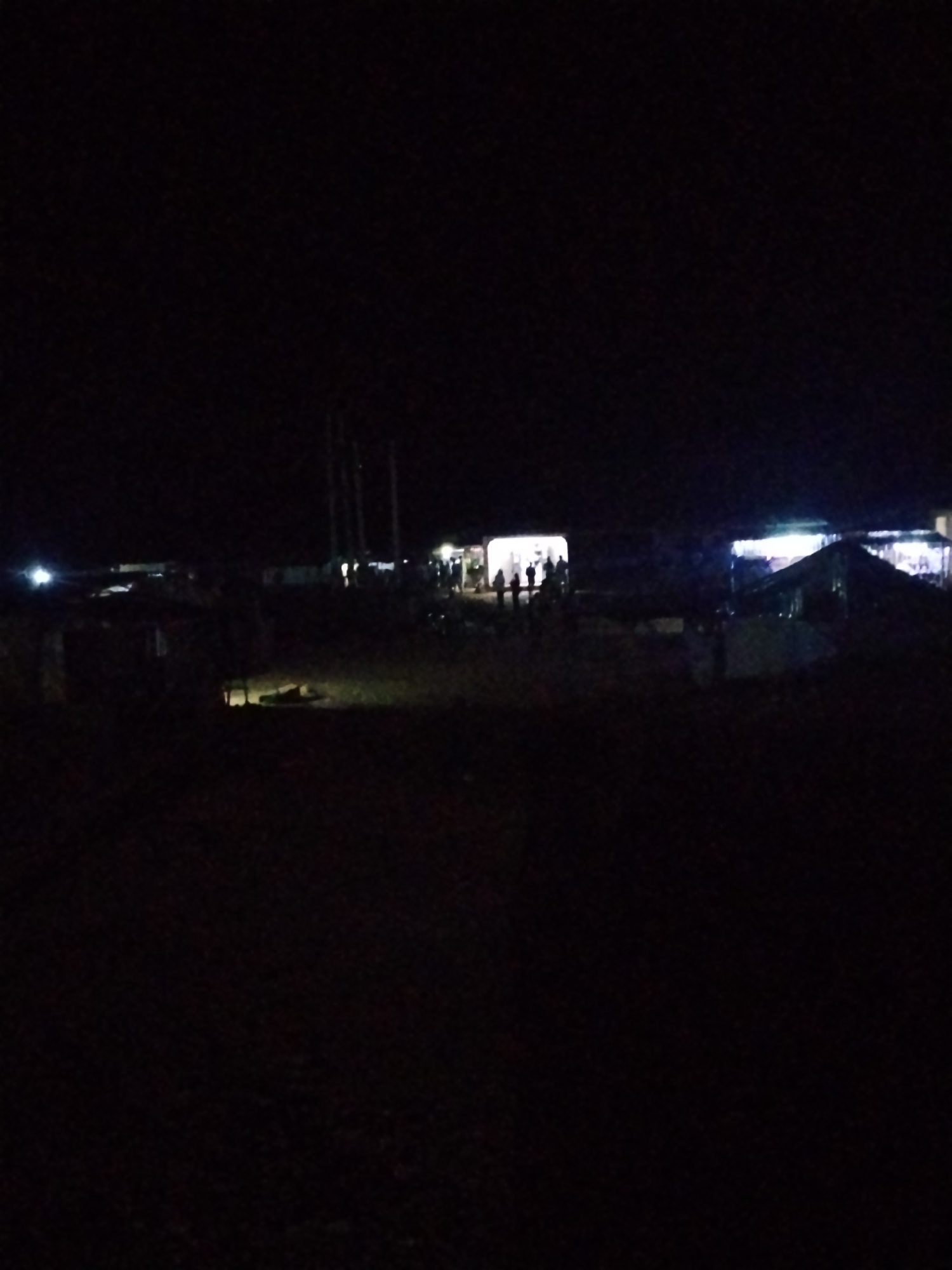 The image depicts a nighttime scene in the city of Durumi, Nigeria, likely an outdoor market or gathering area. The foreground is dark and indistinct, suggesting unlit terrain. In the mid-ground, several structures are illuminated by artificial lights. One structure, possibly a stall or tent, is brightly lit from within, casting a strong glow and silhouettes of people standing in front of it. To its right, another larger structure, also illuminated, appears to be a tent or canopy. Further to the left, a bright light source is visible, possibly a vehicle headlight or a streetlamp. Silhouettes of several individuals are discernible in the lit areas, suggesting a gathering or activity. The overall impression is of a lively, albeit dimly lit, nocturnal community hub.
