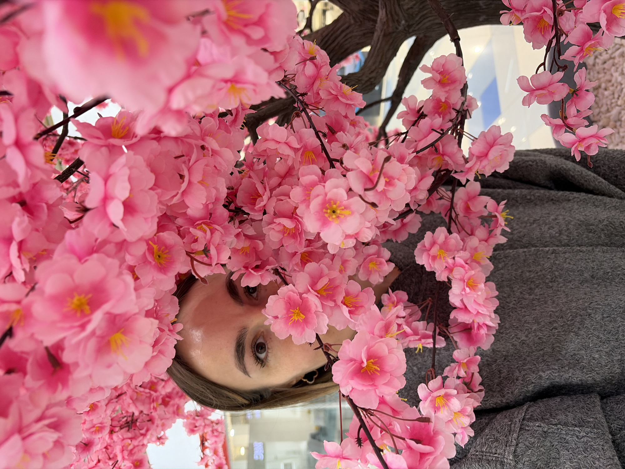 The image shows a person with their face partially obscured by pink cherry blossom-like flowers. The person has light skin, visible eyebrows, and an eye looking towards the camera. They are wearing a gray garment. The background is blurred, but suggests an indoor environment with light and reflective surfaces. The photo appears to be taken from a low angle, looking upwards towards the flower arrangement and the person. It is not possible to confirm the location as Izhevsk, Russia from the image.