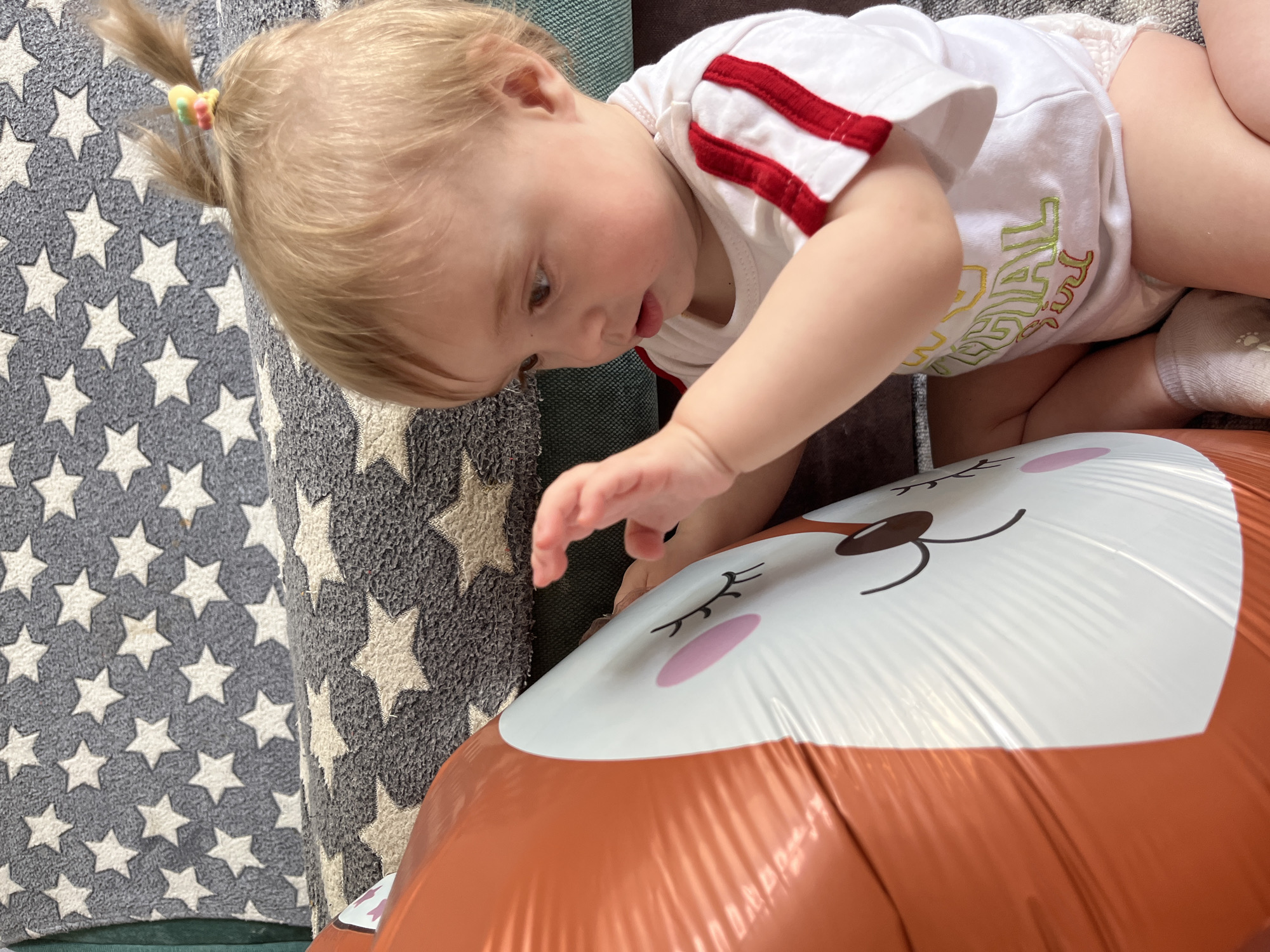 A blonde toddler with a small ponytail held by a colorful hair tie is touching a large, brown-and-white fox-shaped balloon. The child is wearing a white onesie with red stripes on the sleeves and colorful text on the front, and white socks. The child is lying on a patterned gray and white star-covered rug or blanket, which appears to be on a green couch. The scene takes place indoors. There is no information in the image to confirm the location as Chaikovsky, Russia.