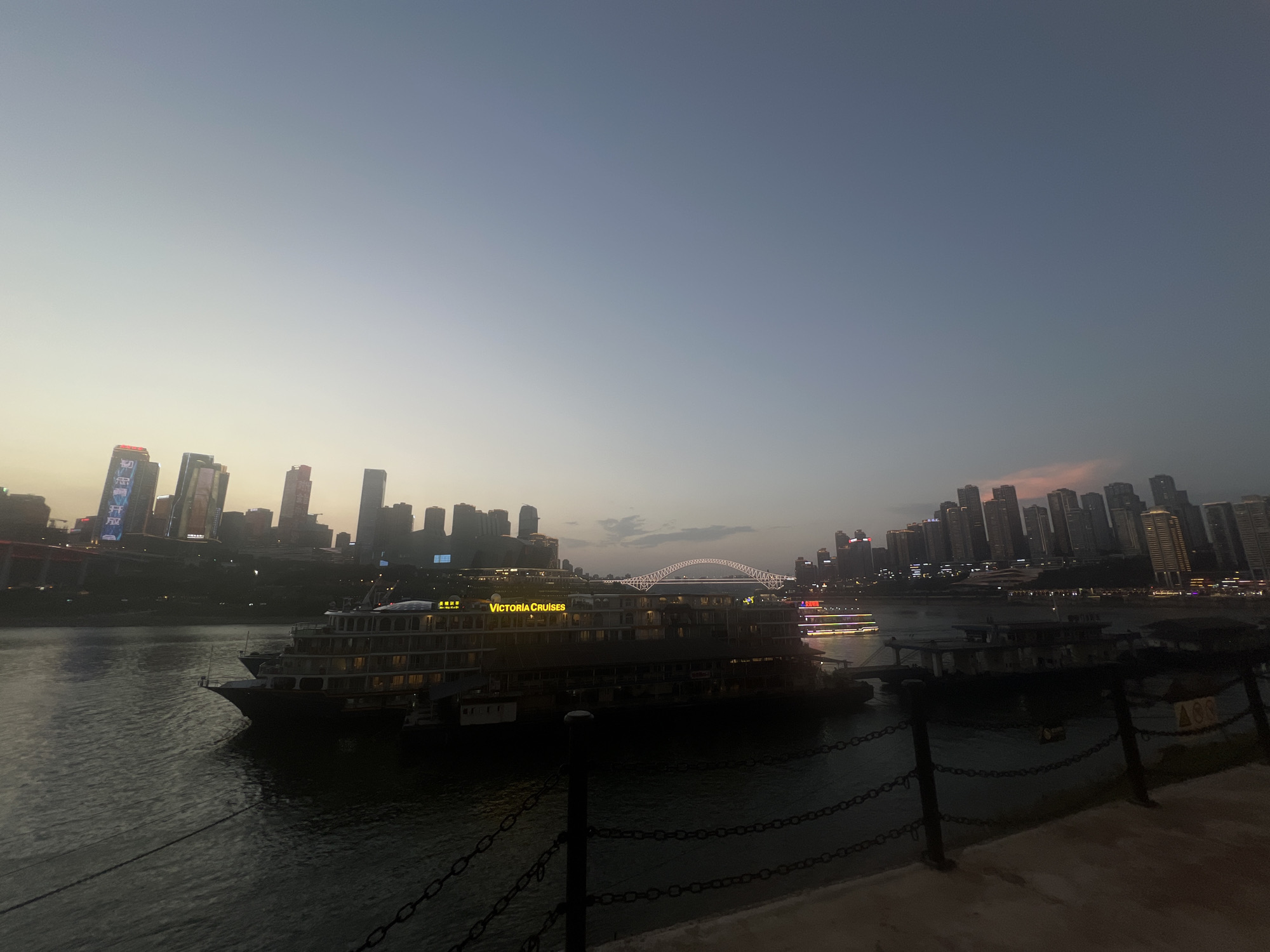 A wide shot captures a dusky cityscape on a riverfront, featuring a large passenger boat named "Victoria Cruises" illuminated with golden lights. The boat is moored in the foreground, with its multiple decks clearly visible, and appears to be the main subject of the scene. In the background, a dense cluster of illuminated skyscrapers pierces the twilight sky, indicating a bustling urban environment. A distinctive arched bridge spans the river in the mid-ground, further emphasizing the city's infrastructure. The water is dark and reflects some of the distant lights. The sky is a gradient of pale blue and soft orange, suggesting either dawn or dusk. The scene is set in Yuzhong District, China, a location known for its dramatic riverfront and mountainous topography, which is hinted at by the tiered arrangement of the city.