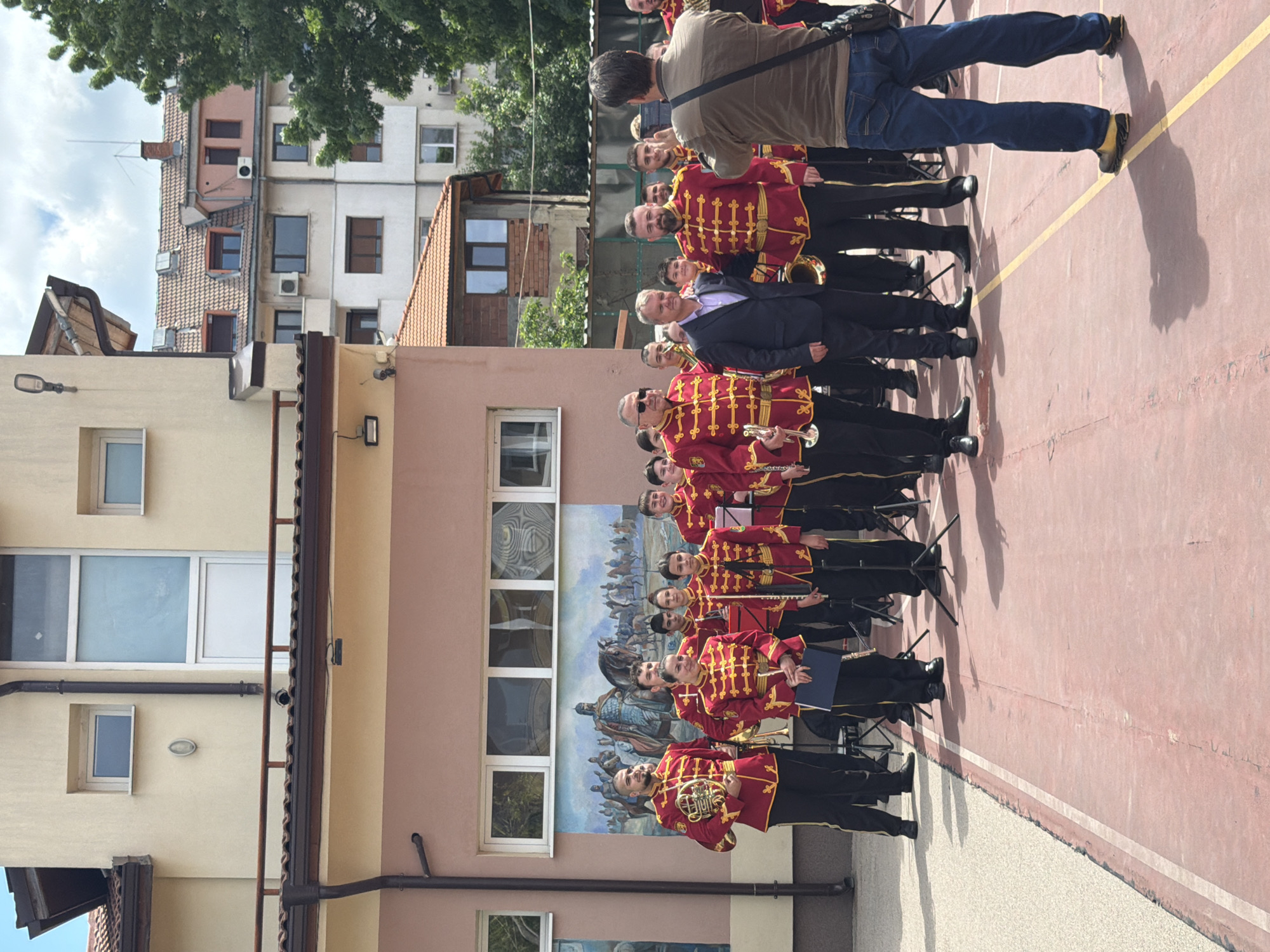 The image shows a brass band standing in formation on a basketball court in Pleven, Bulgaria. The band members are wearing red and gold uniforms with black pants and shoes. An older man in a black suit stands amongst them. A photographer takes a photo. The location appears to be in front of a school, based on the mural and the architecture. Buildings are in the background.