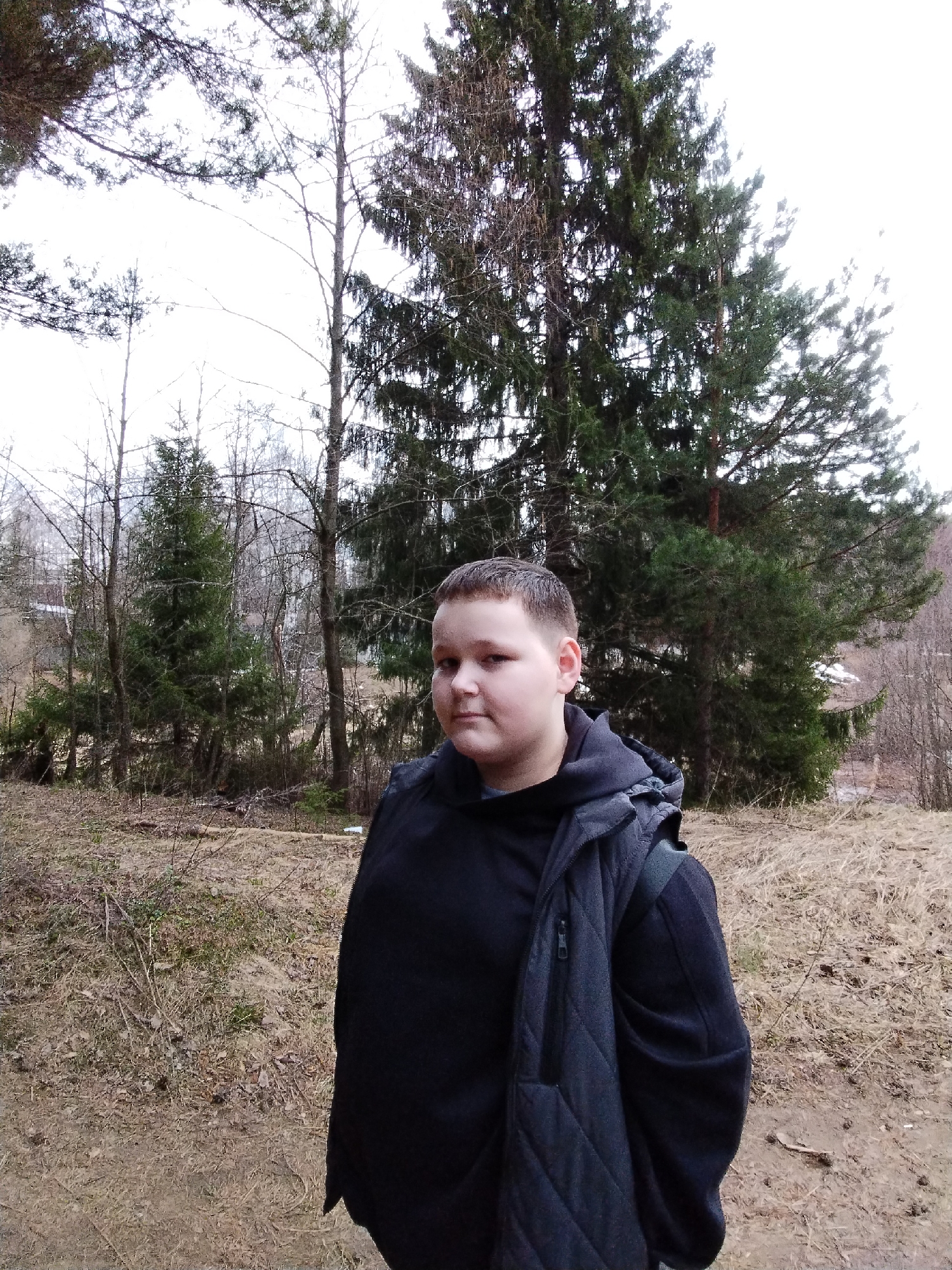 A medium-level, eye-level outdoor shot depicts a young person standing in a forested area. The individual is wearing a black hooded sweatshirt layered with a dark-colored quilted vest or jacket. In the background are various trees, including evergreens and bare trees without leaves, suggesting either late autumn, winter, or early spring. The ground is covered with dry grass, leaves, and dirt. The sky is overcast, creating soft and muted lighting. The subject appears to be in a natural area or park, possibly on the outskirts of or within a city. Given the query's reference to the city of Chaikovsky, Russia, it is presumed that this image was taken in or near a park in Chaikovsky. No specific objects or actions are evident in the scene. The focus is primarily on the individual and the surrounding natural environment.