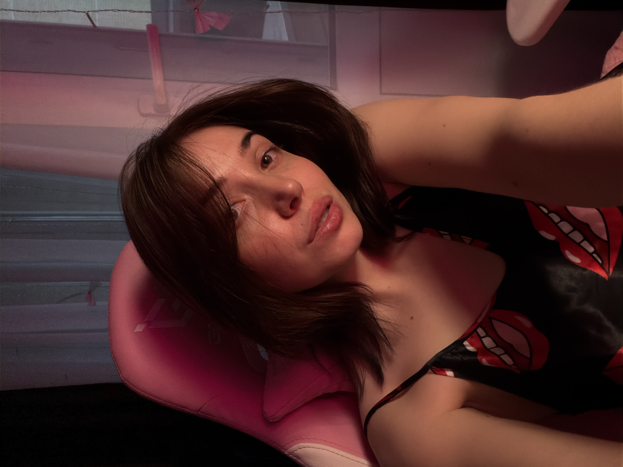 A young woman with brown hair and fair skin, wearing a black slip with a red lips print, is seated in a pink gaming chair. Her head is tilted to her right, and she is looking directly at the camera. The lighting in the room is dim and appears to be from a pink-hued source, casting shadows on her face and body. She is in an indoor setting, with a dark curtain on the left and a window with sheer curtains on the right. The overall mood is intimate and possibly staged. The image does not contain any information about its location in a city or any specific geographical context.