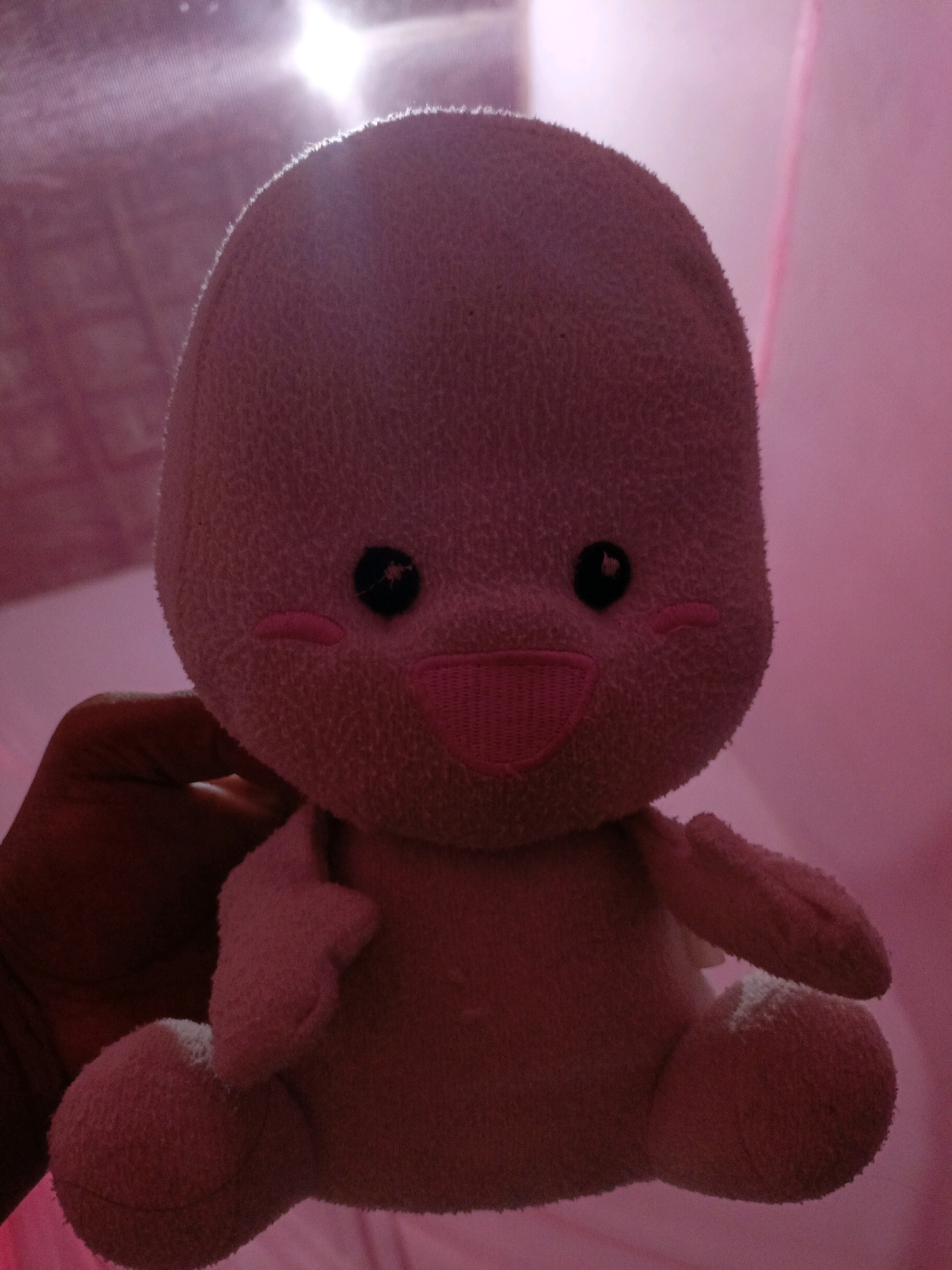 A person's hand holds a pink, fluffy stuffed toy in front of a pink, translucent fabric, possibly a mosquito net or curtain. The stuffed toy has a rounded head with two black button eyes, pink blush marks on its cheeks, and a wide pink stitched smile. It has small, stubby arms and legs. A bright light source is visible in the upper left background, casting a glow and creating highlights on the toy's head. The overall lighting is dim and pink-toned. The image provides no information about the location in the city of Jenu, Indonesia.