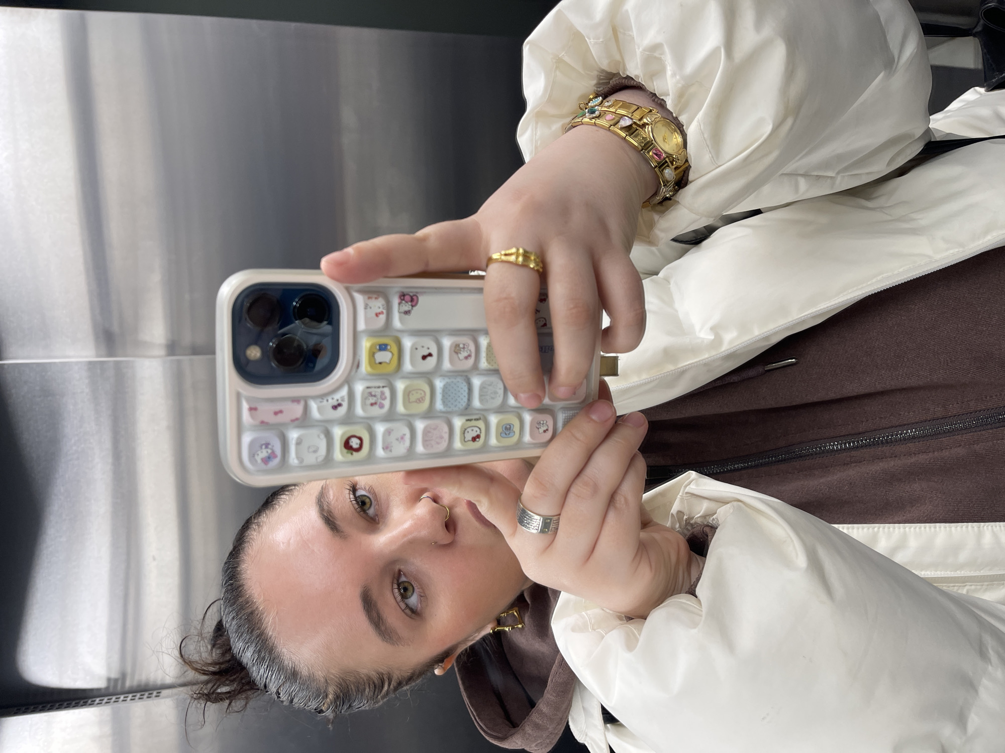 A selfie captures a person, likely female, reflected in a metallic surface, suggesting an elevator or a modern interior. The image is rotated 90 degrees clockwise. The subject has light skin, green eyes, and dark hair pulled back. She wears a septum ring, a nostril piercing, and gold-colored, geometric earrings.
She holds a smartphone, which is partially visible, featuring a unique white case with a grid of colorful square buttons depicting various cartoon characters, including Hello Kitty. The phone's multi-lens camera system is prominent.
Her left hand, holding the phone, displays a thin gold-colored ring on the ring finger and a silver-colored ring with an engraved pattern on the middle finger. Her right wrist is adorned with a striking gold-colored watch with a round face and a band embellished with colorful gems and pearls.
The person is dressed in an off-white or cream-colored puffy jacket, worn over a dark brown zipped hoodie or top. Visible zippers are present on both garments. The background is a reflective metallic panel, which reflects light and parts of the subject. The person's expression is neutral as she looks towards the camera, capturing her reflection. The location context provided is Moscow, Russia, though the image itself does not offer specific geographical cues.