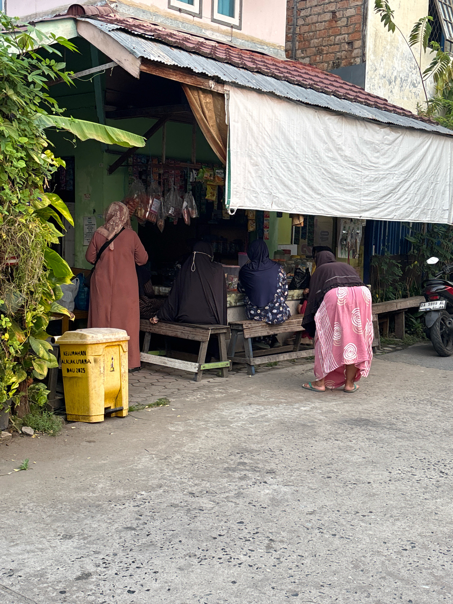 This image depicts a street scene in Banjarmasin, Indonesia, likely near a residential area or small commercial hub, with a focus on a small roadside store. In the foreground, a yellow trash can with text indicating "KELURAHAN ALALAK UTARA DAU 2025" is visible on the cracked concrete pavement. Behind it, a lush green plant with large leaves cascades down the left side.
The store itself has a green facade and a corrugated metal roof supported by wooden beams. A white fabric awning extends over the front of the store, obscuring some of the view. Hanging from the store's interior are various packaged goods, suggesting it sells snacks or small grocery items.
Several individuals are present, all appearing to be women dressed in traditional Islamic modest wear, including hijabs. One woman stands near the trash can, facing the store, wearing a long, rust-colored robe and a patterned hijab. Another woman, in a black hijab and robe, is seated on a wooden bench in front of the store. Next to her, a woman in a dark blue hijab and floral-patterned dress is also seated on the bench. A fourth woman, wearing a dark head covering and a long, flowing pink skirt with a white circular pattern, is bent over near the bench, seemingly interacting with something on the ground or at the store.
A red motorcycle with license plate "DA 5999 AL" is parked to the right of the scene, further indicating a street-side setting. The environment suggests a warm climate, with the presence of foliage and the casual attire of the people. The overall atmosphere appears to be that of a typical neighborhood store where people gather.