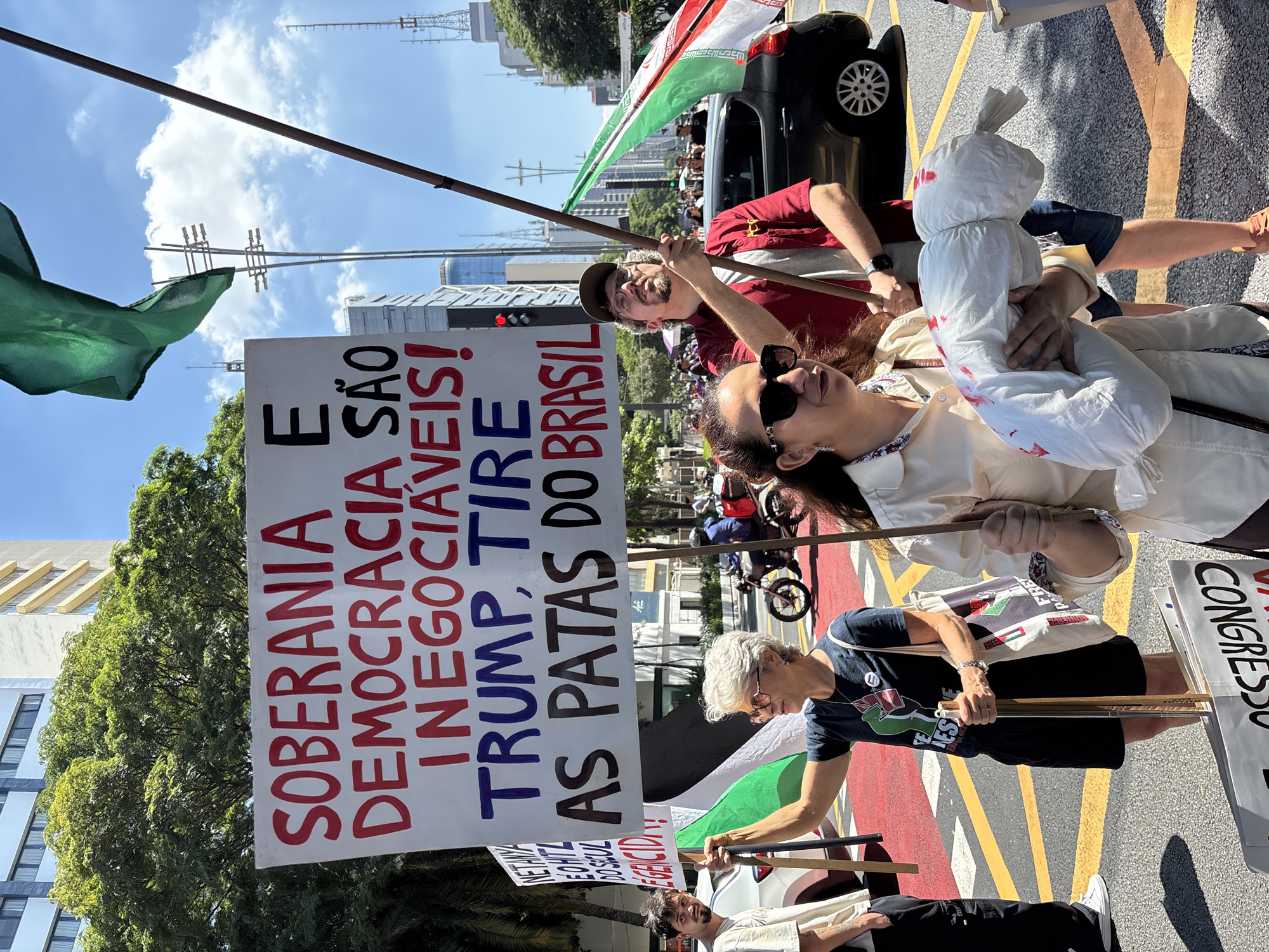 A street-level, eye-level shot depicts a political demonstration in what appears to be a city environment, likely in São Paulo, Brazil. The image is slightly tilted.
Several individuals are visible, carrying signs and flags. A large, rectangular sign, prominently positioned, displays text in Portuguese, with the phrase "SOBERANIA E DEMOCRACIA SÃO INEGOCIÁVEIS! TRUMP, TIRE AS PATAS DO BRASIL" dominating the sign.
People are holding flags; a green and white flag is visible. One individual carries what appears to be a effigy wrapped in white material.
The setting shows a street marked with yellow and red lines, typical of urban traffic lanes. In the background, buildings, trees, and utility poles suggest a city. A black car can be seen in the background.