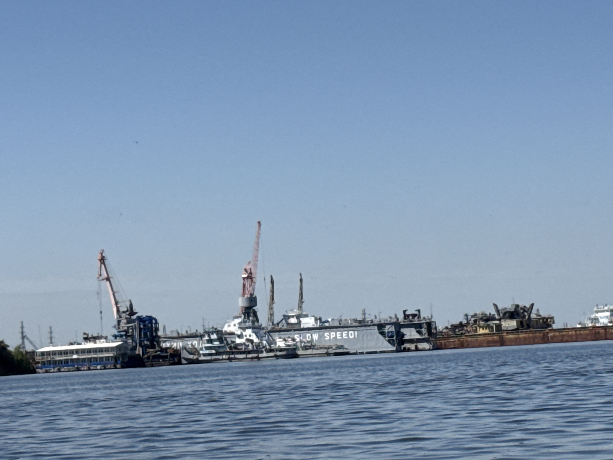 The image depicts a waterfront scene, possibly near a shipyard or industrial area, under a clear, bright sky. The water surface is rippled, indicating slight movement. Several vessels are visible, including barges and ships, some of which are docked or moored. One barge prominently displays the words "SLOW SPEED!" in large letters. Cranes are positioned near the vessels, suggesting ongoing construction, repair, or loading/unloading activities. Given that the city of Chaikovsky is specified, the scene likely represents an industrial area along the Kama River. The overall impression is one of an active, potentially working waterfront.