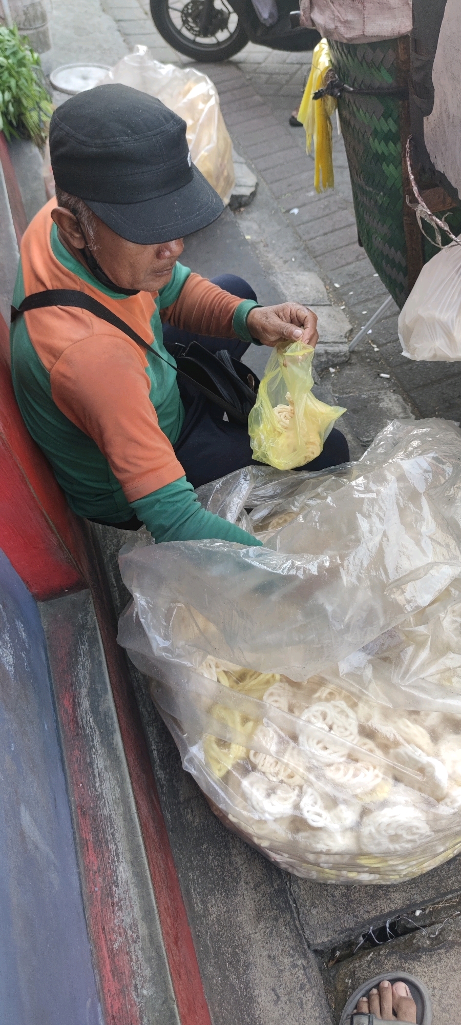 An elderly man, wearing a black baseball cap, an orange and green long-sleeved shirt, and dark pants, sits on a low, red and grey concrete ledge. He holds a small, translucent yellow plastic bag filled with coiled, light-colored food items, likely snacks. Before him, on the ground, is a large, translucent plastic bag overflowing with similar coiled food items, appearing to be crackers or crisps. The man is looking down at the bag he is holding. To his right, a portion of a green and brown woven basket is visible, attached to what appears to be a cart or display. A portion of a motorcycle wheel is visible in the background. The ground is paved with grey concrete blocks. The scene is set on a street in Sruni, Indonesia.