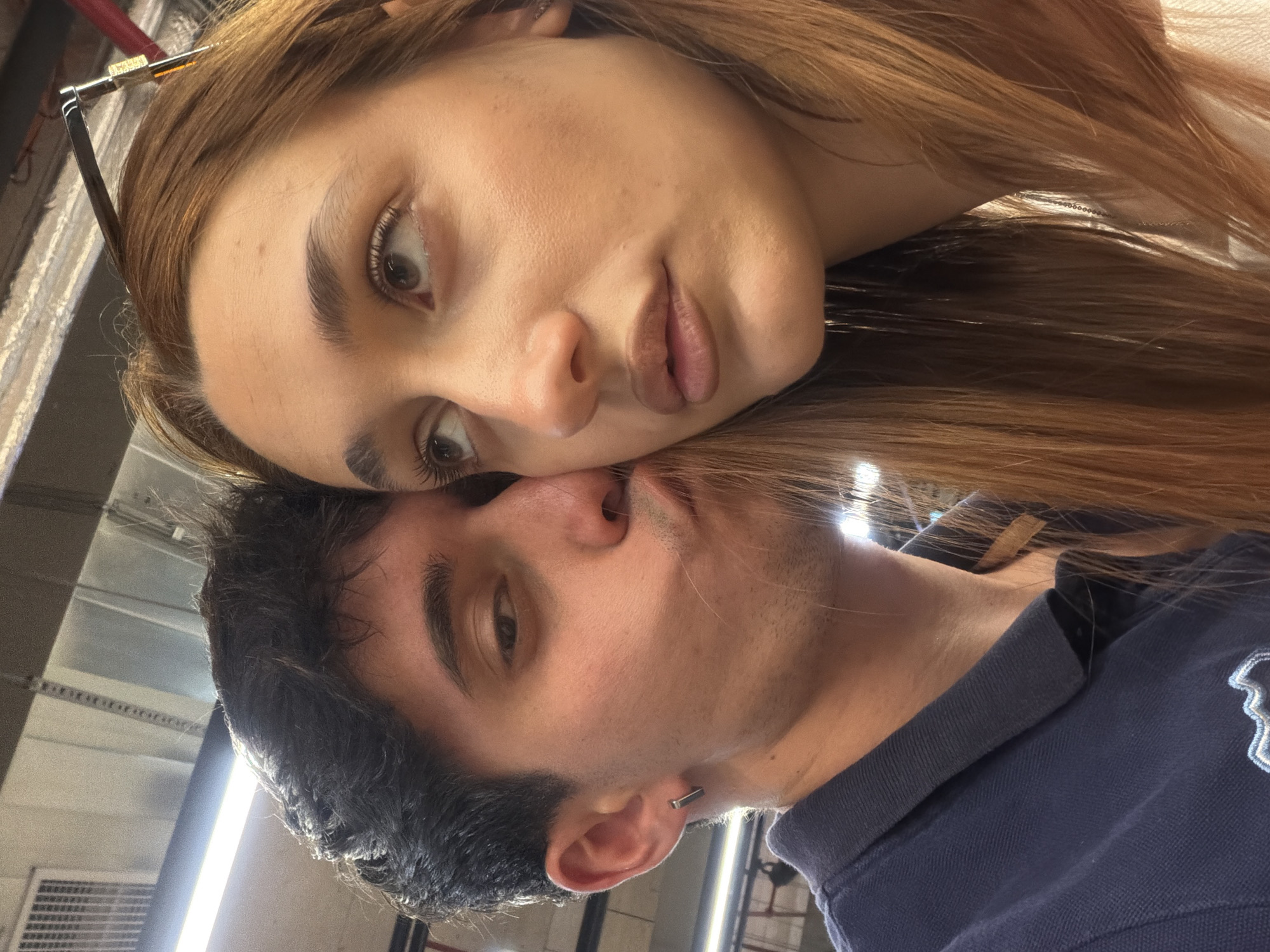 A medium-shot, indoor, selfie image shows a young woman lying her head on top of a young man's head. The young woman has fair skin, and brown hair, and is wearing glasses. The young man has dark hair, fair skin and a dark navy polo shirt. They appear to be located in a metro station, likely in the city of Sofia, Bulgaria.