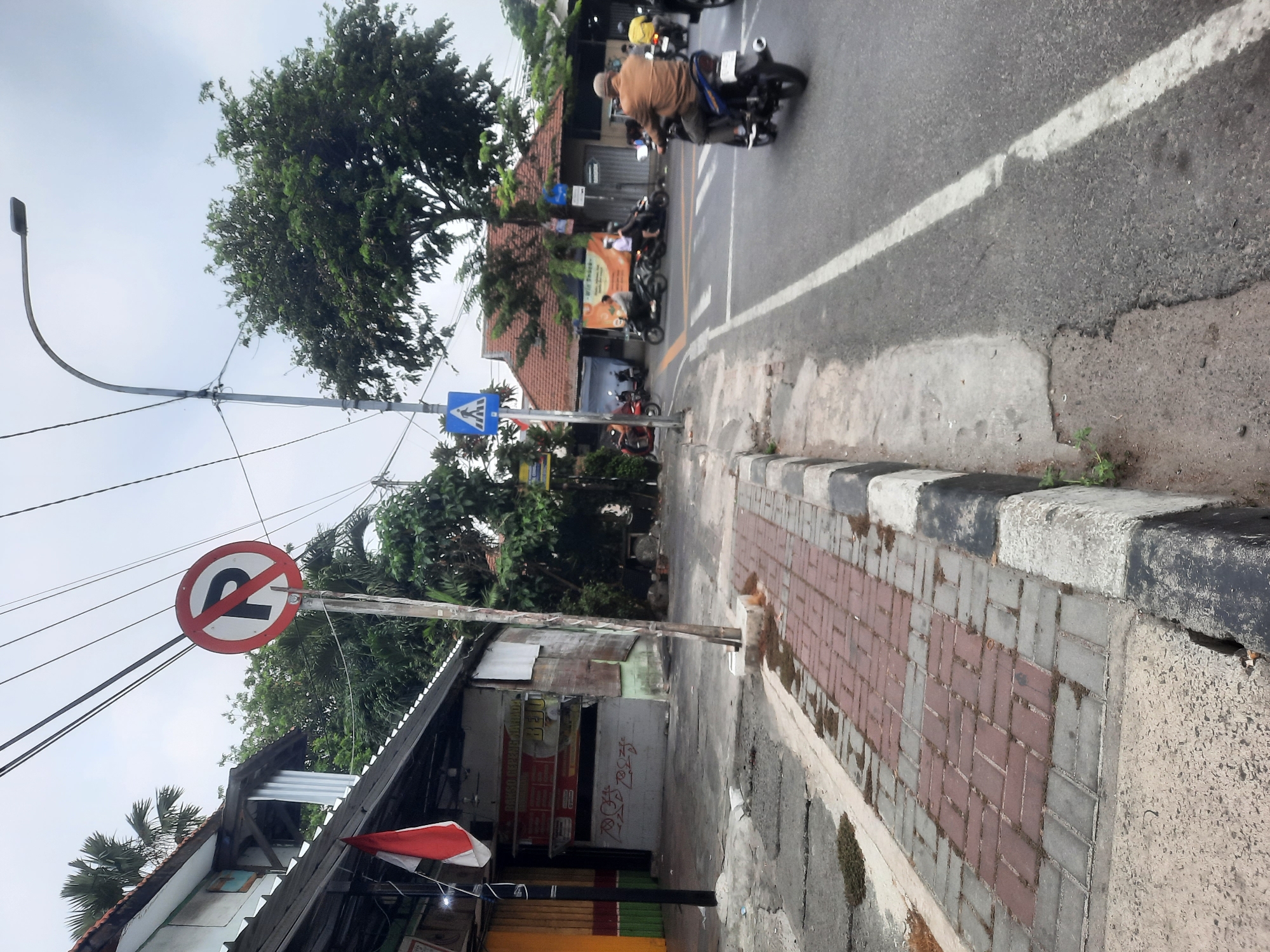 The image depicts a street scene in North Jakarta, Indonesia, on an overcast day. A paved road runs diagonally from the bottom right towards the upper left, with a white line marking the lane. Several motorcycles are visible on the road; one in the foreground is ridden by a man wearing a light brown jacket and a cap, another is ridden by a person in a white shirt, and further back, a red motorcycle is seen.
To the left of the road is a sidewalk with a brick-patterned surface and a black and white striped curb. Adjacent to the sidewalk is a building with a gray and white facade and a dark-colored roof. A red and white Indonesian flag is displayed on the left side of this building. Another building with a red-tiled roof is visible behind the first building.
Several traffic signs are present. A circular sign with a red border and a red slash over a black "P" indicates "no parking." A blue square sign with a white pedestrian symbol denotes a crosswalk. A street lamp with a curved arm extends over the scene. Trees with dense green foliage are scattered throughout the background. Electrical wires are strung across the sky. The overall impression is of a typical urban street environment with moderate pedestrian and vehicle activity.