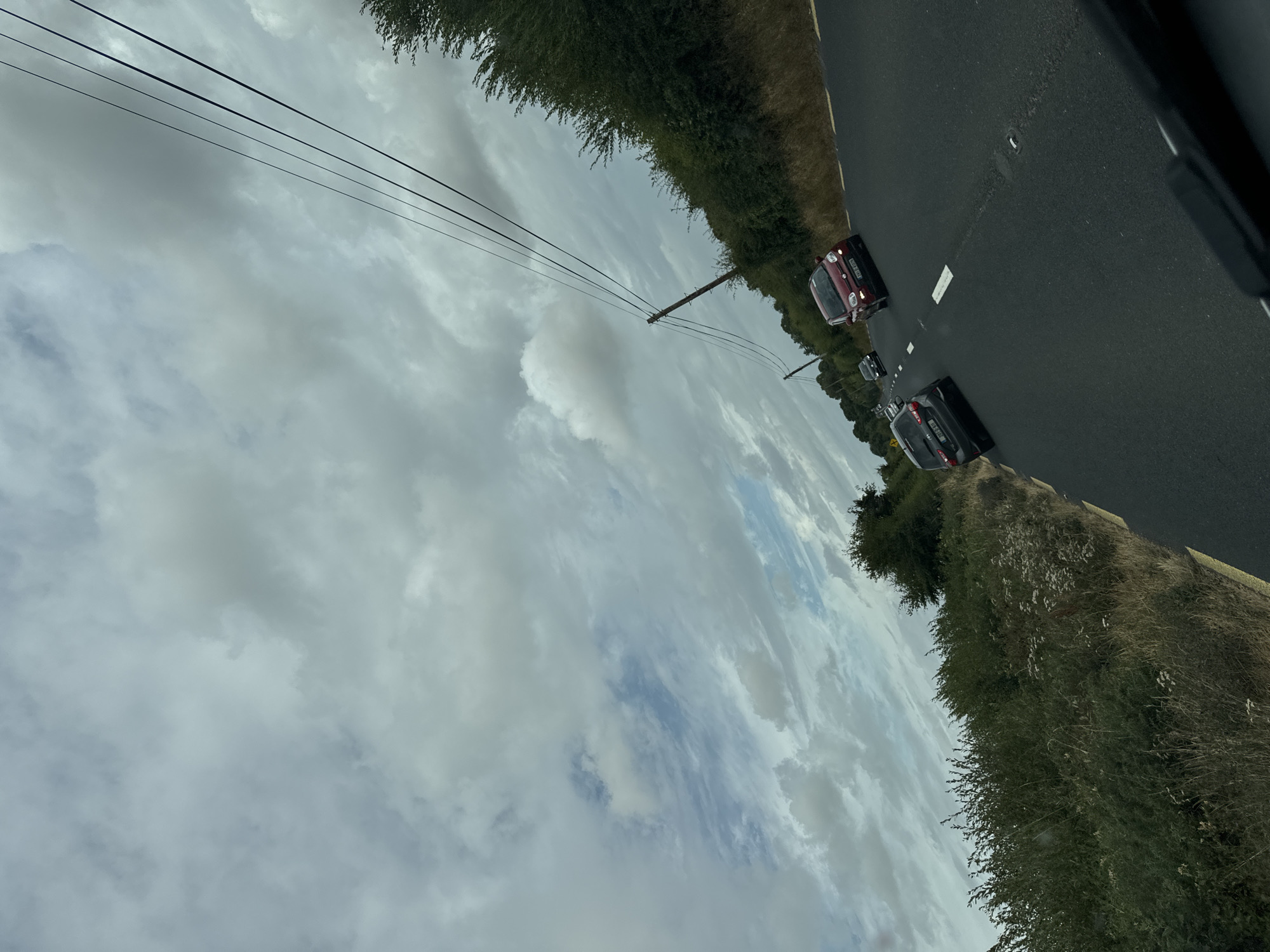 The image shows a view from a vehicle driving on a rural road in New Ross Municipal District, Ireland. The sky is filled with heavy, gray clouds, with patches of blue sky visible. Three cars are on the road ahead, a dark gray hatchback in the left lane, followed by a red hatchback in the right lane. A fourth car, a darker color, is visible further ahead. The road is asphalt, with a yellow line marking the edge on the left and a dashed white line separating the lanes. Lush green vegetation, including bushes and trees, lines the right side of the road, with taller trees in the distance. Utility poles with overhead wires run along the right side of the road. The scene appears to be during daylight hours, likely overcast.