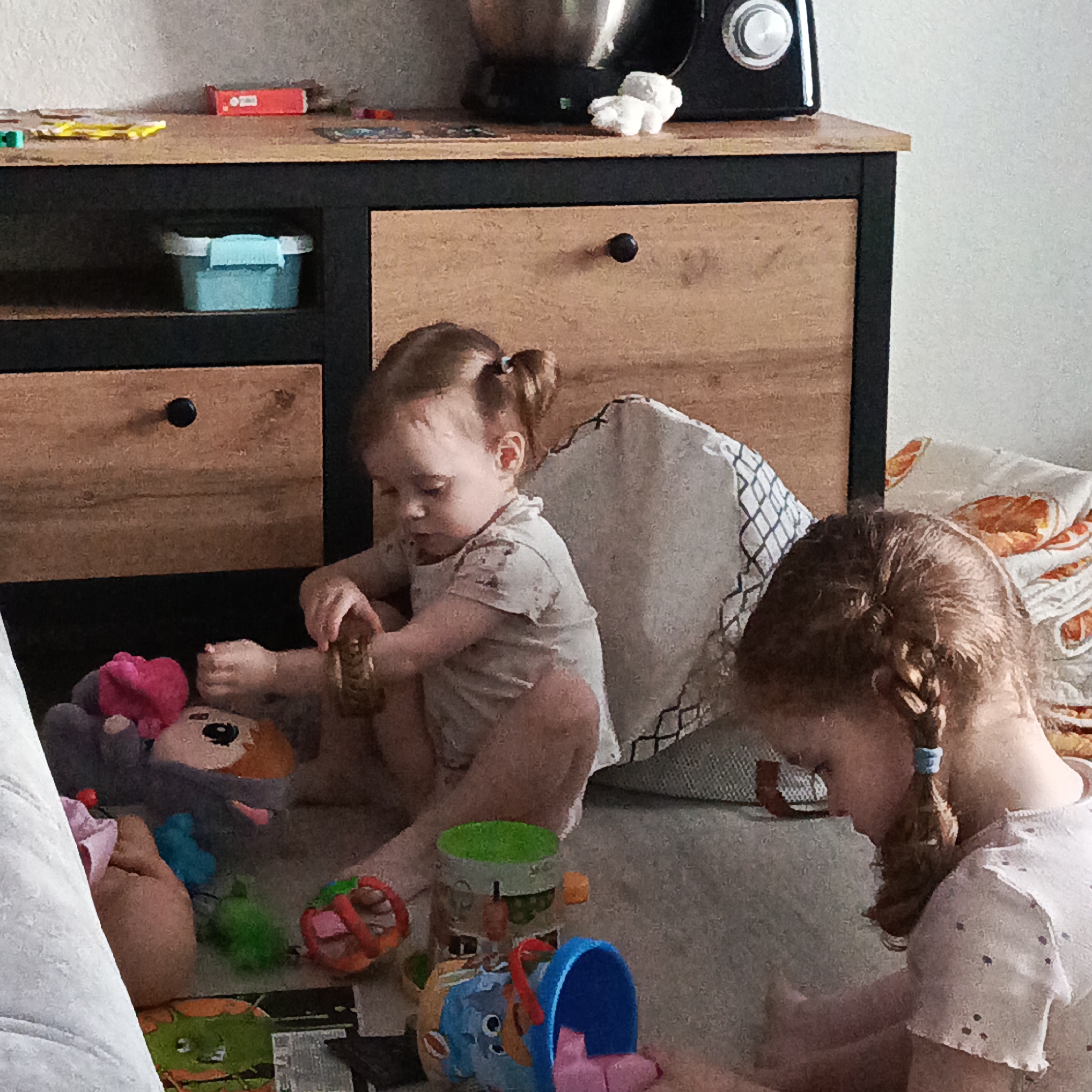 In a domestic setting in Марковский, Russia, two young girls are playing on the floor in front of a wooden cabinet. The cabinet has a black frame, wooden drawer fronts, and black round knobs. A kitchen appliance sits on top of the cabinet. A baby girl with her hair in pigtails is sitting and holding a toy. An older girl with her hair in braids is sitting and looking down. The floor is covered with toys including stuffed animals and plastic containers. Pillows are scattered behind the girls.