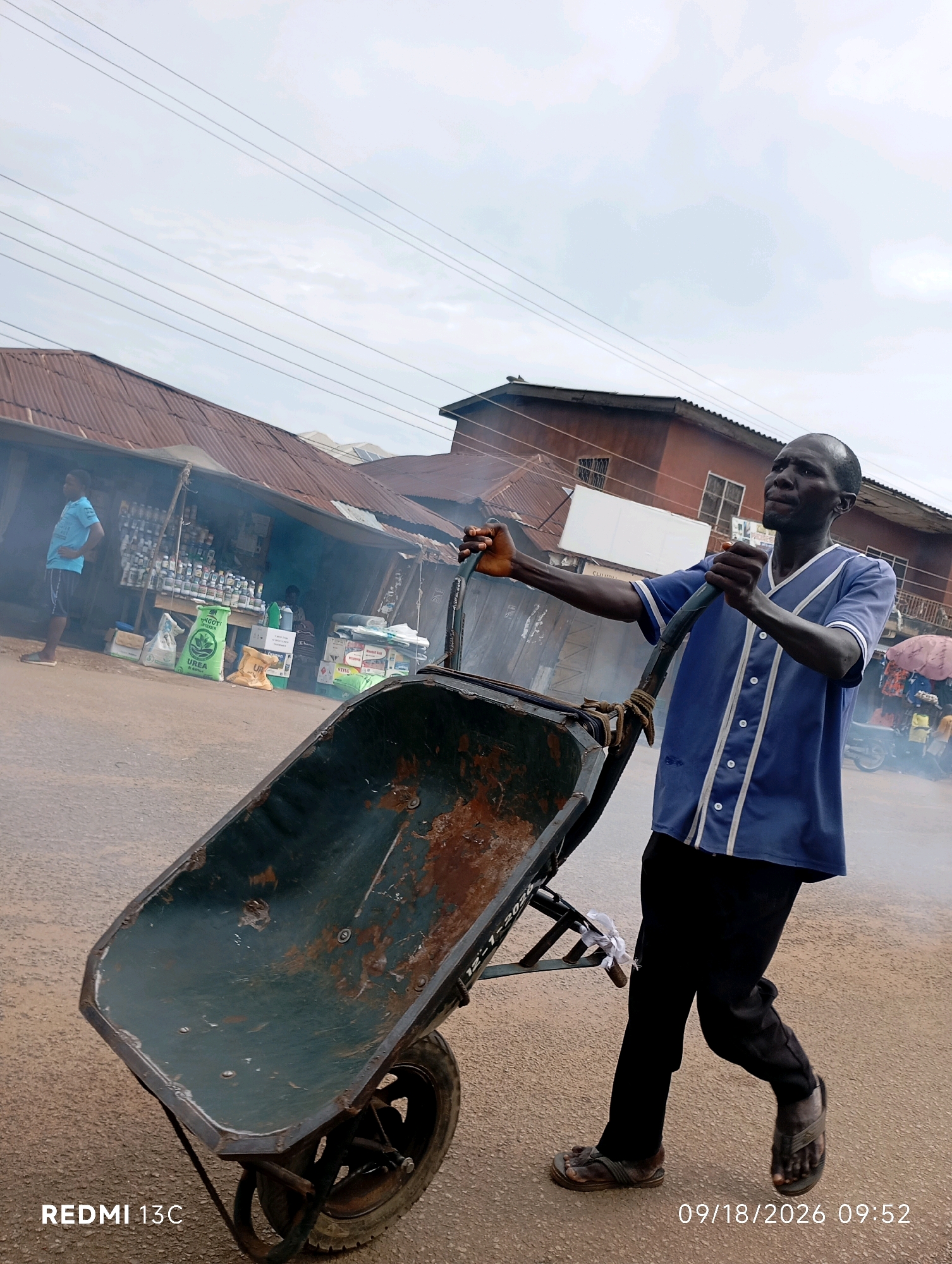 In Shendam, Nigeria, a man in a blue and white baseball jersey and black pants pushes a rusty, dark green wheelbarrow down a dusty, unpaved road. Smoke emanates from the left side of the frame, obscuring a stall with various goods displayed, including sacks of fertilizer and bottles. Another man, wearing a blue t-shirt and shorts, stands in front of the stall. The background shows a row of buildings with corrugated iron roofs, typical of a market area. The sky is overcast. The timestamp "09/18/2026 09:52" and "REDMI 13C" are visible in the lower right and left corners, respectively.