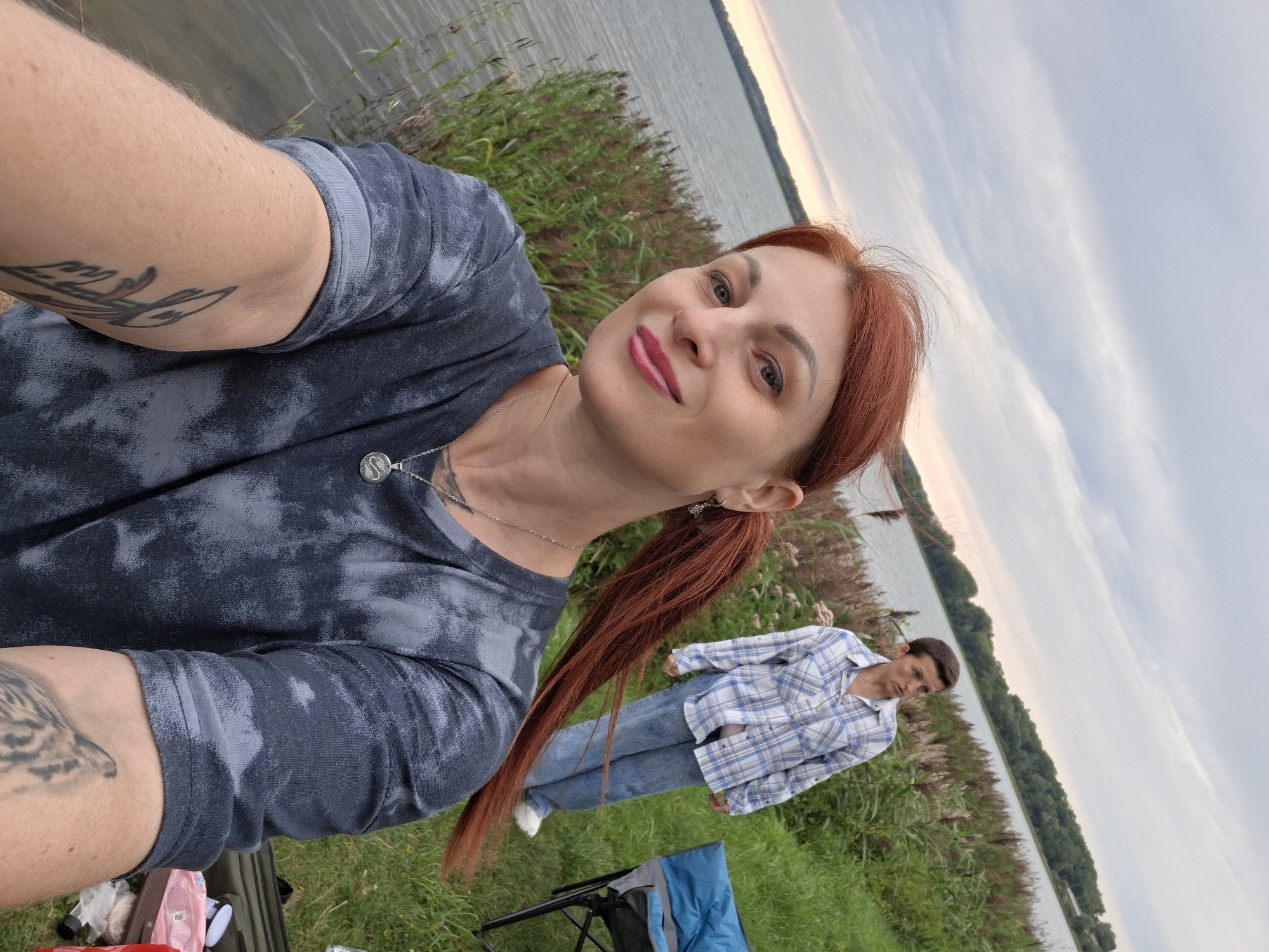 In this image, a woman with long, red hair pulled into a ponytail is taking a selfie. She is wearing a dark, tie-dye t-shirt and a silver necklace with a circular pendant. Tattoos are visible on her arm. In the background, a second person is standing, wearing a blue and white checkered shirt and jeans. They are on a grassy area near a body of water, with tall reeds and trees in the distance. The sky is overcast. This scene is consistent with a location on the shores of Lake Myastro in Myadzyel, Belarus.