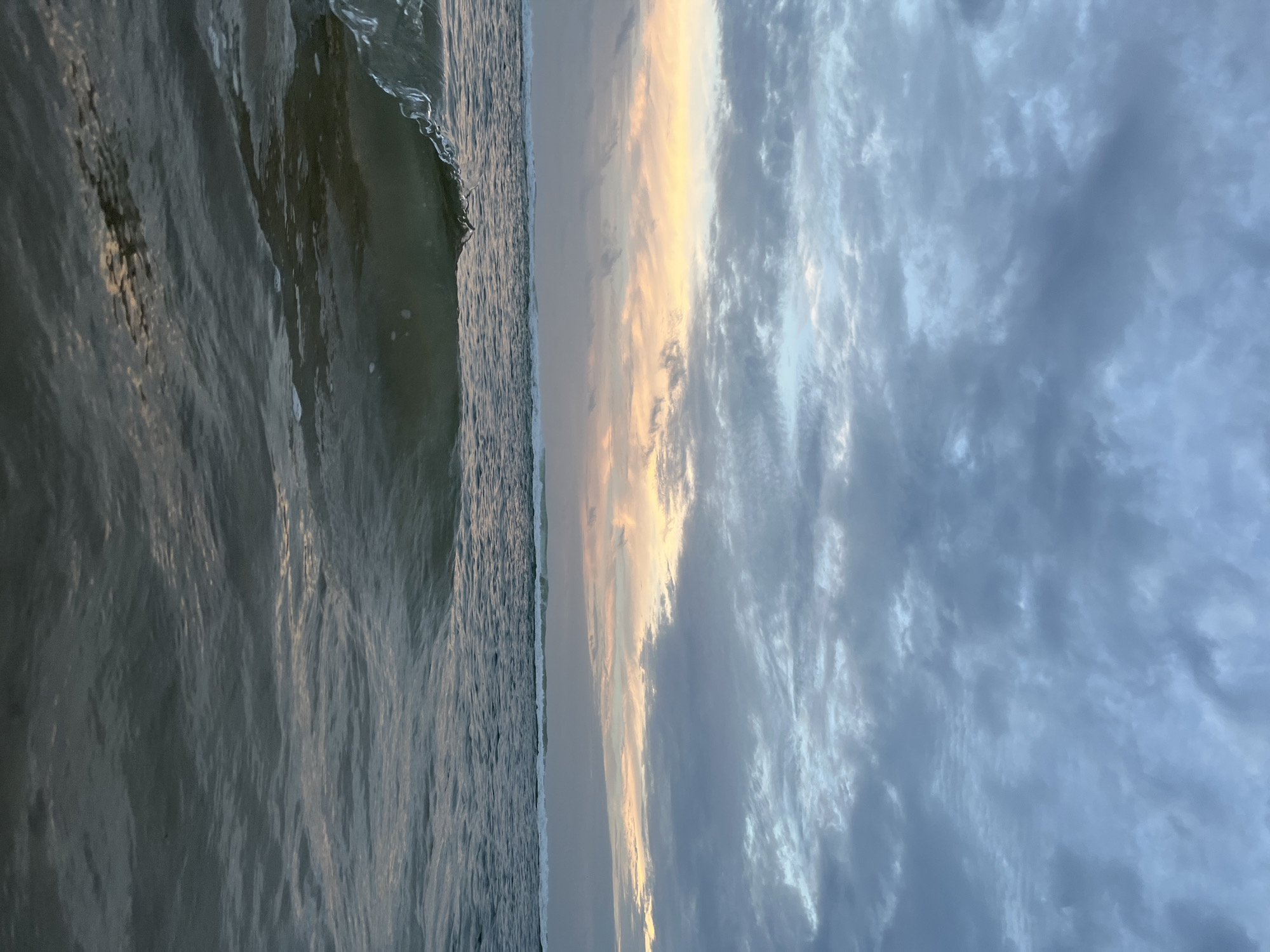 The image depicts a serene seascape at dusk, taken from a low angle, possibly from within the water. The left side of the frame is dominated by the ocean, with gentle waves reflecting the muted light of the setting sun. The water appears dark and somewhat choppy, with subtle ripples creating a textured surface. A small wave crests on the far left, its translucent edge catching the light.
The right side of the image is dedicated to the sky, which is a canvas of dramatic clouds painted with the warm hues of sunset. A band of golden-yellow light emanates from the horizon, illuminating the undersides of scattered clouds and creating a striking contrast with the deeper blues and grays of the sky above. The clouds are dense and textured, suggesting a dynamic weather pattern.
The scene evokes a sense of calm and natural beauty, with the transition from day to night being the central theme. The time of day is clearly evening, as indicated by the sunset colors. The location cues, though not explicitly visible in the image, are identified as Kathaluwa, Sri Lanka, suggesting a tropical coastal setting. There is no visible text in the image. The overall mood is peaceful and contemplative, capturing the fleeting moment of sunset over the ocean.