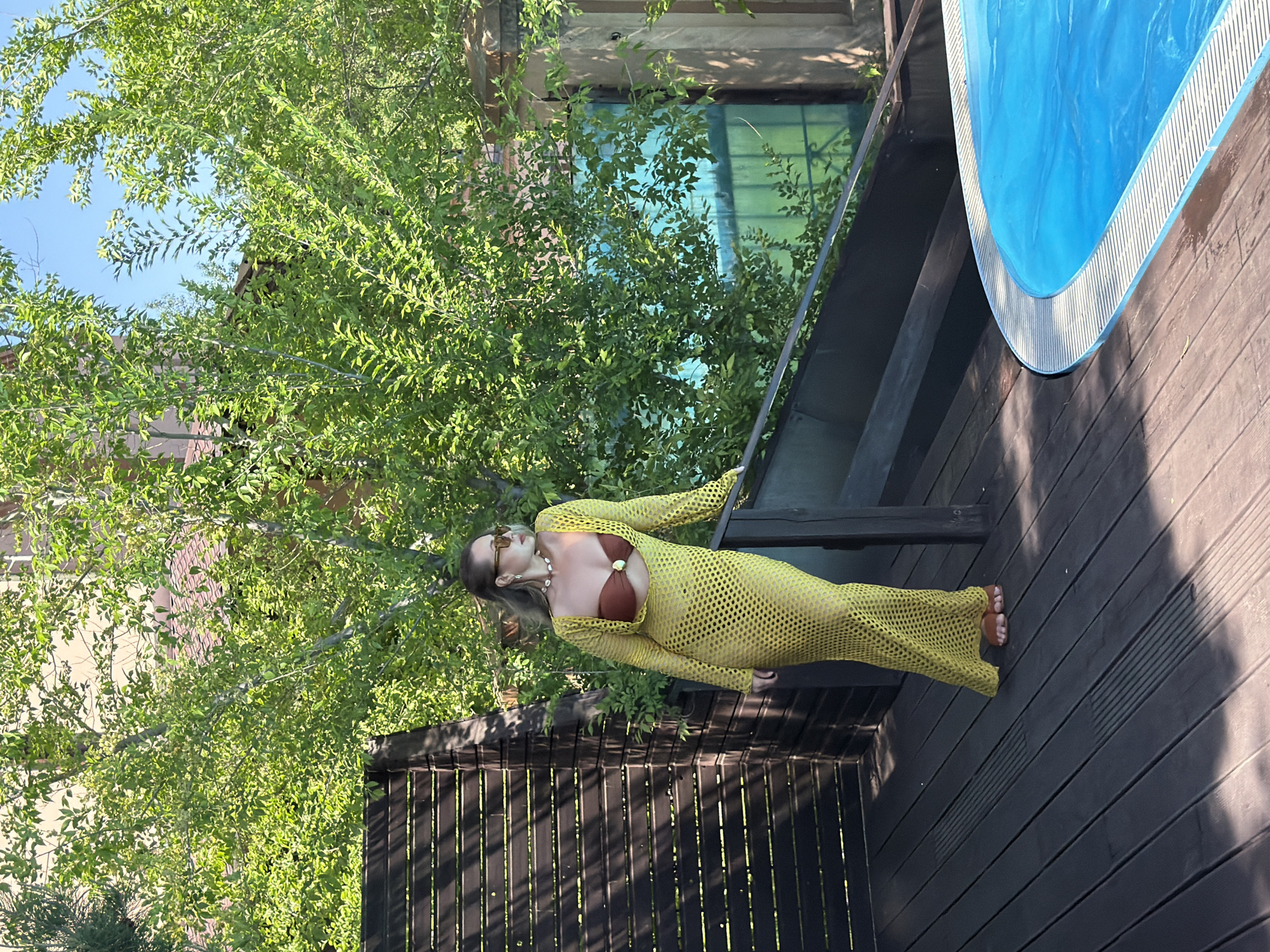 This media file, when correctly oriented, presents an outdoor scene on a bright, sunny day. The central subject is a woman standing on a dark wooden deck. She has light-colored hair and is wearing sunglasses, a long-sleeved, yellow, open-knit mesh cover-up over a brown bikini top, and a necklace featuring white, shell-like beads. Her left hand rests on a dark wooden railing, and her bare feet are visible on the deck. She appears to be posing for the camera, exhibiting a neutral expression.
To the right of the woman, a blue swimming pool with a white patterned edge is partially visible, suggesting a recreational area. The background is dominated by lush, vibrant green trees and dense foliage, indicating a natural or well-landscaped setting. Through the tree canopy, portions of a building with a reddish-beige facade and windows, possibly reflecting the sky or surrounding greenery, can be glimpsed. Dark wooden fences or additional railings are also present within the scene. The strong sunlight casts clear shadows, signifying midday or early afternoon. The overall atmosphere is summery and relaxed. There is no visible text within the image. The context for this location is Krasnoyarsk, Russia.