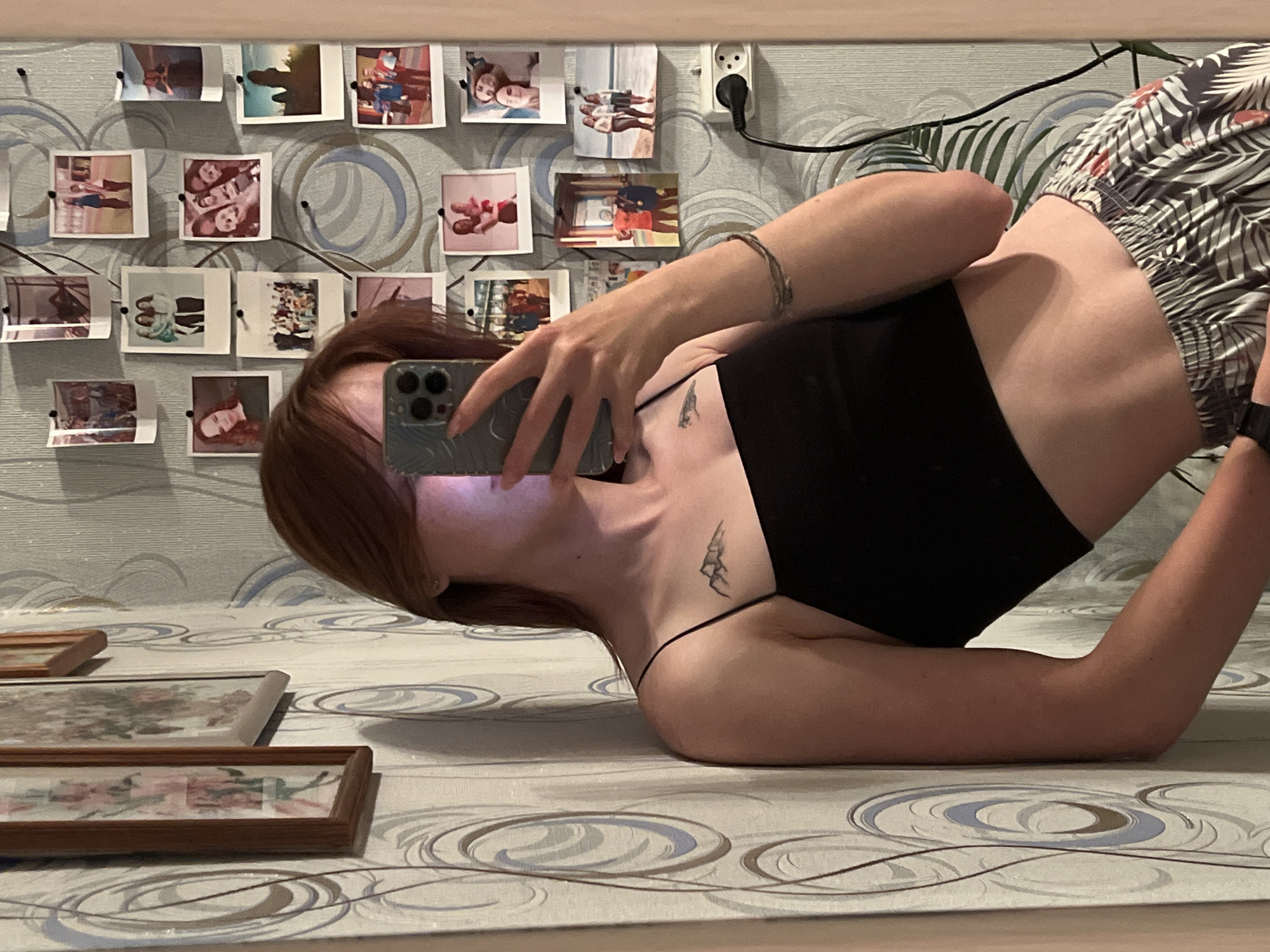 The image shows a young woman taking a selfie in a room, leaning against a wall with patterned wallpaper. She is wearing a black spaghetti strap crop top and patterned shorts. There are framed pictures and small polaroid photos pinned to the wall behind her. A power outlet is visible on the right side of the wall. The woman has a tattoo on her upper chest. The overall impression is of a casual, personal space, likely a bedroom. There is no information in the image that would allow for the identification of its location in Chaikovsky, Russia.