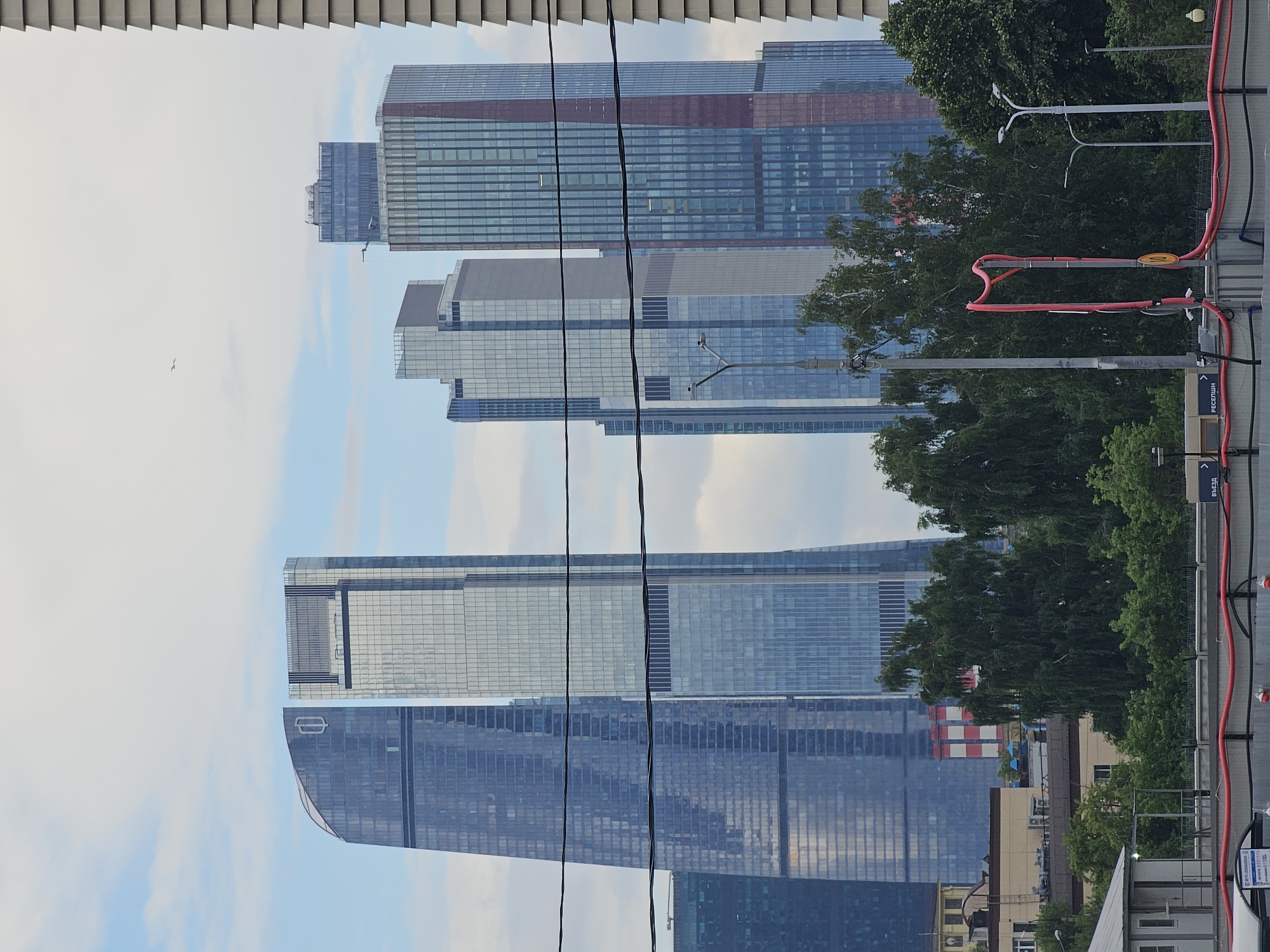 This image shows a view of modern skyscrapers in Moscow, Russia, on a cloudy day. The skyscrapers are tall, glass-fronted buildings with distinct architectural designs. In the foreground, there are power lines and some trees. The image is oriented vertically, with the sky at the top and the ground at the bottom. The overall impression is one of urban development and a bustling city.
The most prominent subjects are the skyscrapers. They appear to be office buildings or residential towers, with many windows and varying heights. Some of the buildings have a reddish-brown hue at the top, suggesting a different material or color scheme. In the middle ground, there are large trees with lush green foliage, which contrast with the sharp lines of the architecture. The sky is overcast with some visible clouds, indicating a temperate but not necessarily sunny day.
There are no people visible in the image. The activity taking place is essentially the passive observation of the cityscape. The time of day appears to be daytime, likely mid-morning or afternoon, given the ambient light.
Notable details include the power lines running across the frame, which add a sense of urban infrastructure to the scene. The red cables in the foreground are also a distinct visual element. There is some visible text on signs in the image, but it is mostly illegible due to the distance and resolution. One sign appears to have Cyrillic script, reinforcing the Russian location.
The setting is clearly urban and modern, characteristic of a major metropolis like Moscow. The combination of towering buildings and surrounding greenery suggests a planned urban environment with parks or green spaces interspersed among the developments. The overall mood is one of a contemporary, functional city.