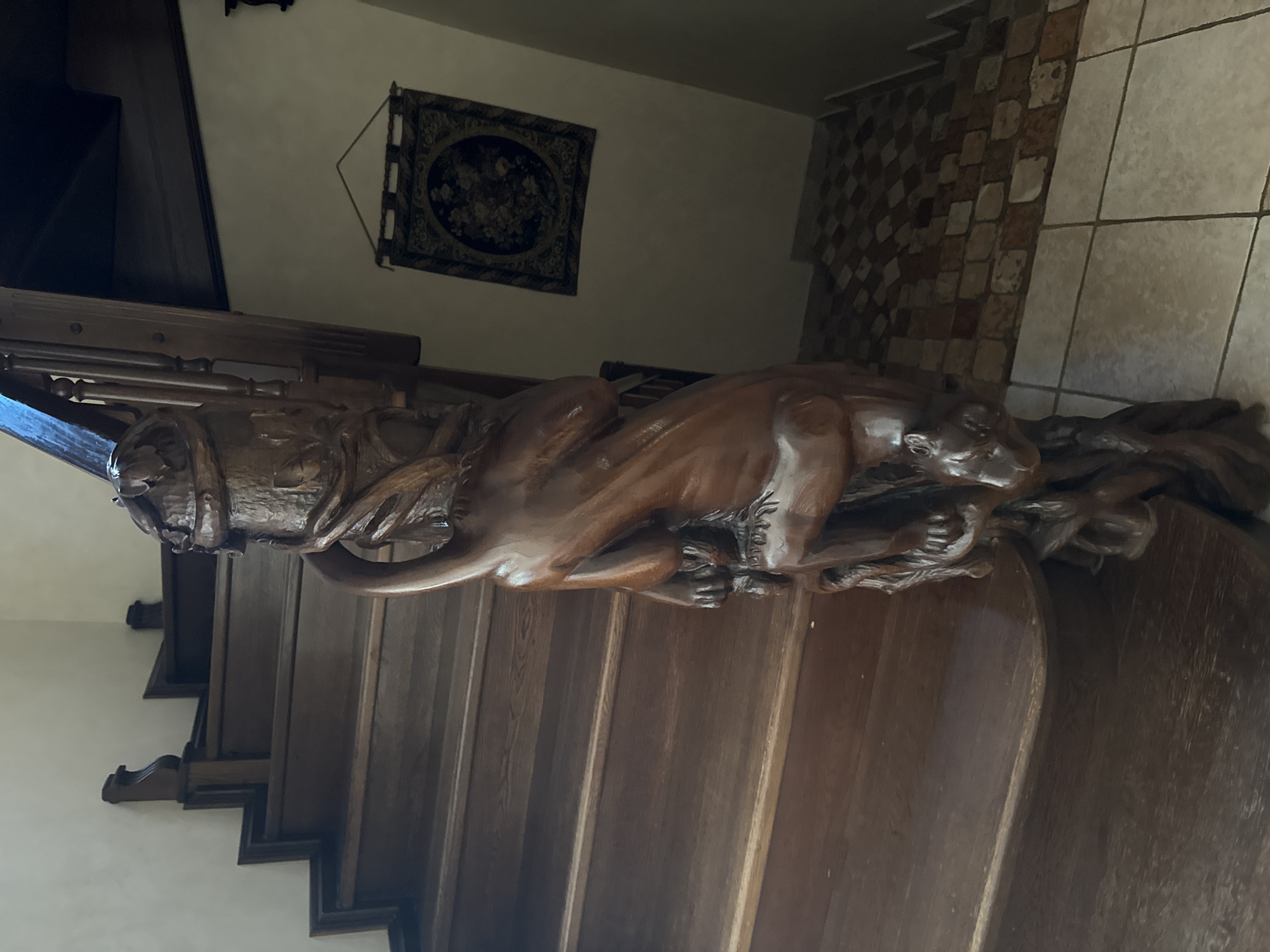 A wooden staircase in a residential building features a carved banister depicting a creature, possibly a dragon or a mythical beast, coiled around a central pole. The creature is intricately detailed, with visible claws, scales, and a head. The staircase itself is made of dark wood and appears well-maintained. The surrounding environment includes a tiled floor at the base of the stairs and a tapestry hanging on the wall. The lighting suggests an indoor setting, likely a home. The location is specified as Yukki, Russia.