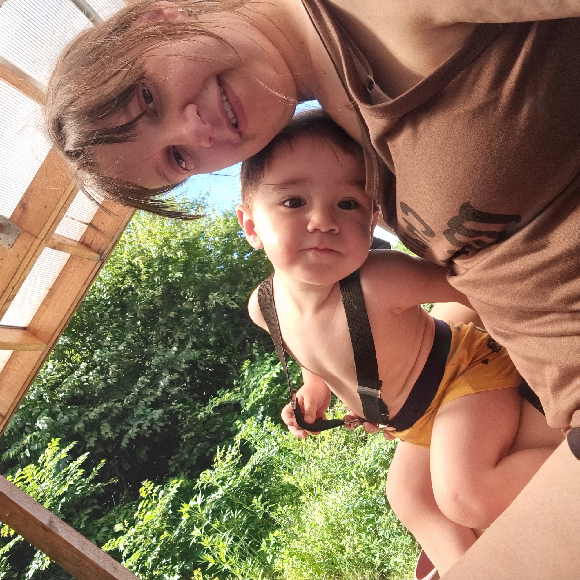 The image depicts a close-up, selfie-style shot of a woman and a baby outdoors, nestled under a wooden and translucent roof structure, likely a pergola or shelter. The background consists of lush green foliage and a clear blue sky, suggesting a warm, sunny day.
The woman, who appears to be in her late 20s or 30s, is positioned in the foreground, looking directly at the camera with a gentle smile. She has light brown hair with bangs and is wearing a brown tank top. Her head is tilted slightly, and her right arm is bent, supporting the baby.
The baby, resting on the woman's chest and shoulder, is facing slightly away from the camera, with its head resting on the woman's collarbone. The baby has dark brown eyes and appears to be very young, likely a toddler. It is wearing yellow shorts with suspenders. The baby's hand is visible, touching the suspenders.
The setting is outdoors, with a focus on the natural environment of trees and greenery. The wooden structure overhead provides a partial frame for the scene. The overall atmosphere is intimate and relaxed, capturing a moment between a mother and child. The specific location in Dunnae-myeon, South Korea, is not discernible from the visual elements in the image.