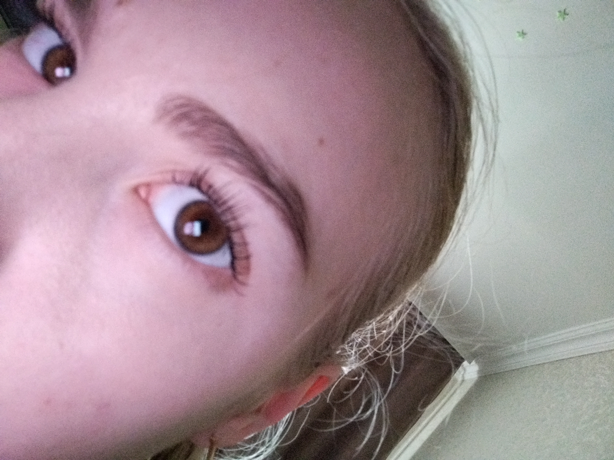 This is a close-up, upward-facing, angled shot of a young person's face. The subject has brown eyes and appears to be looking at the camera. The skin is fair and smooth, with a few faint freckles visible on the forehead and cheek. The eyebrows are well-defined and arched. The hair is light brown and pulled back, with some strands framing the face. The background is out of focus, showing a light-colored wall with decorative molding and some faint, star-shaped stickers. The lighting suggests an indoor setting, possibly daytime. The overall mood is neutral, with no strong emotional cues. There is no visible text in the image.