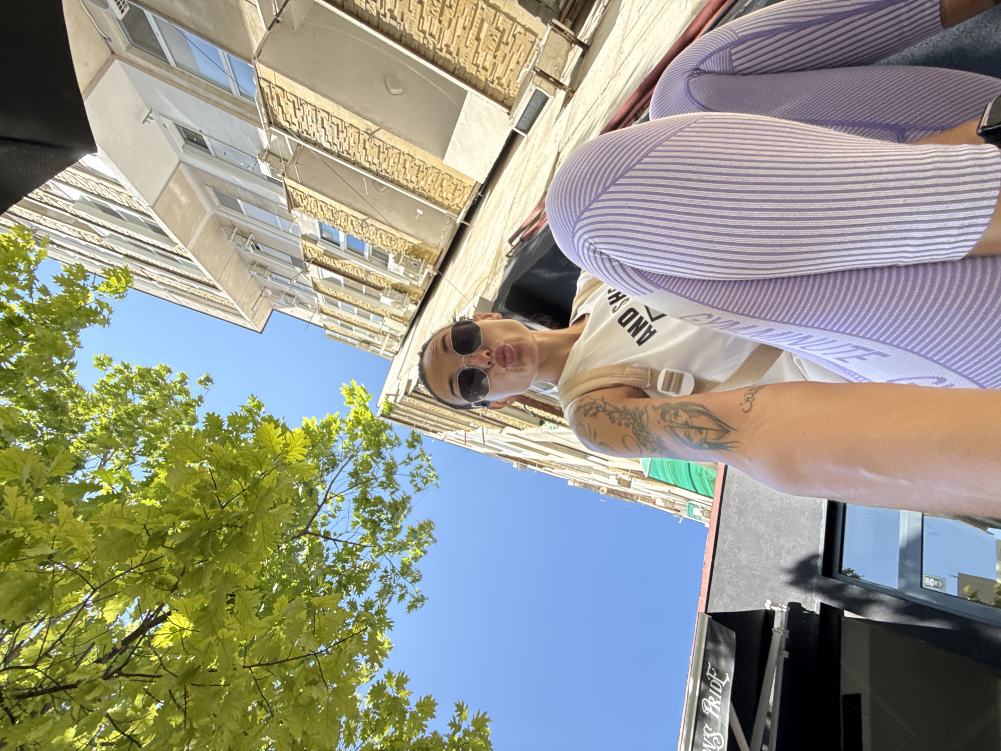 A low-angle, wide-shot POV selfie captures a woman posing in a street in Pleven, Bulgaria. She is wearing a white t-shirt with text, sunglasses, and purple and white striped leggings. She is pouting and holding the camera out. Her arm, adorned with tattoos, is visible in the foreground. To her left, a building with an awning that reads "PRIDE" is visible, along with a tree with green leaves. To her right and behind her, a tall, beige building with many windows and air conditioning units rises into the clear blue sky. The overall scene depicts an urban environment on a sunny day.