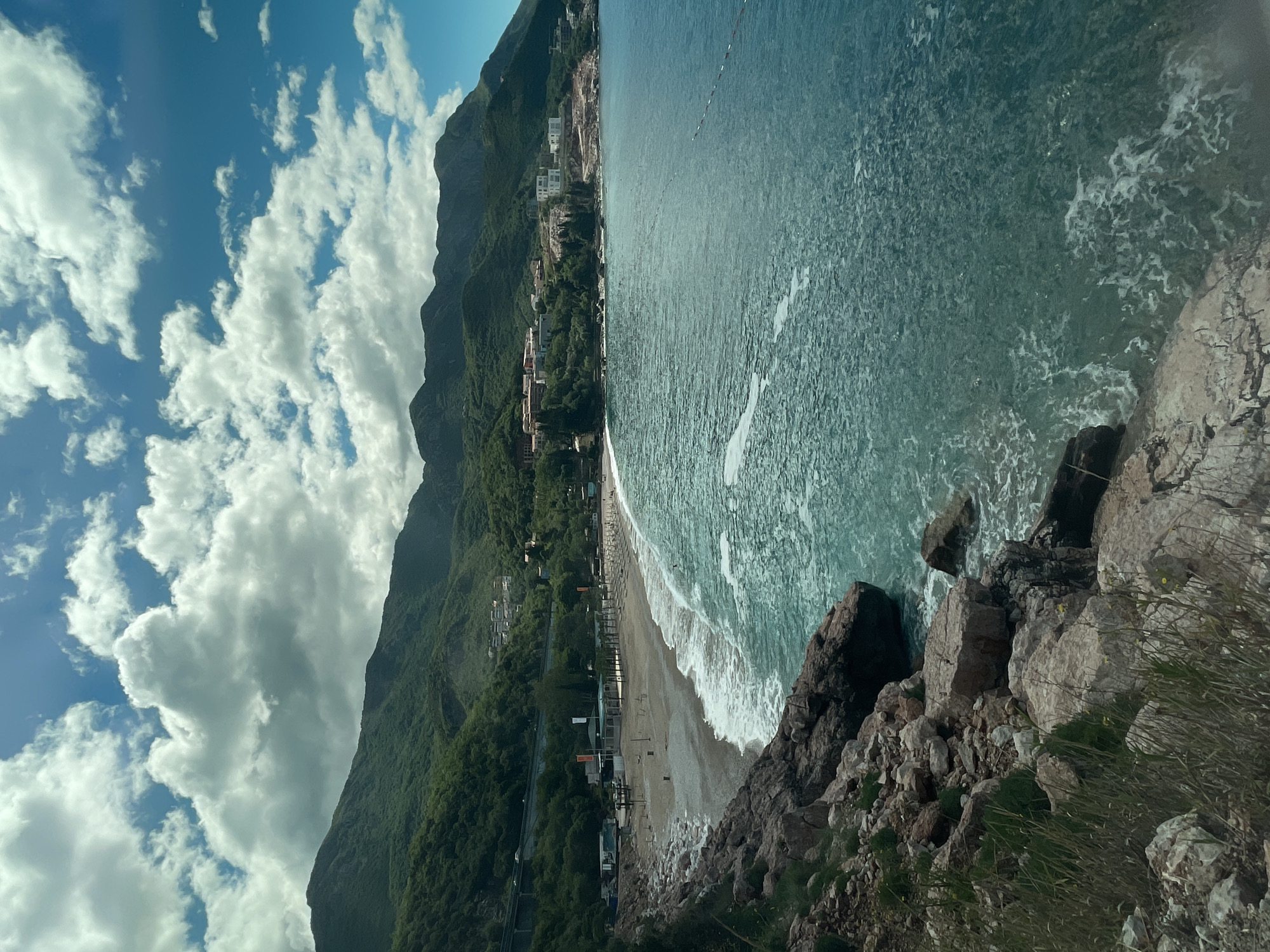 The image captures a coastal scene in what appears to be a mountainous region. The foreground features a rocky cliff edge with some vegetation. Beyond the cliff, a beach curves along the coastline, with gentle waves breaking along the shore. Buildings and structures are visible along the beach and further inland, nestled against the base of a large, green mountain range. The sky is partially cloudy, with a mix of blue sky and white, puffy clouds.