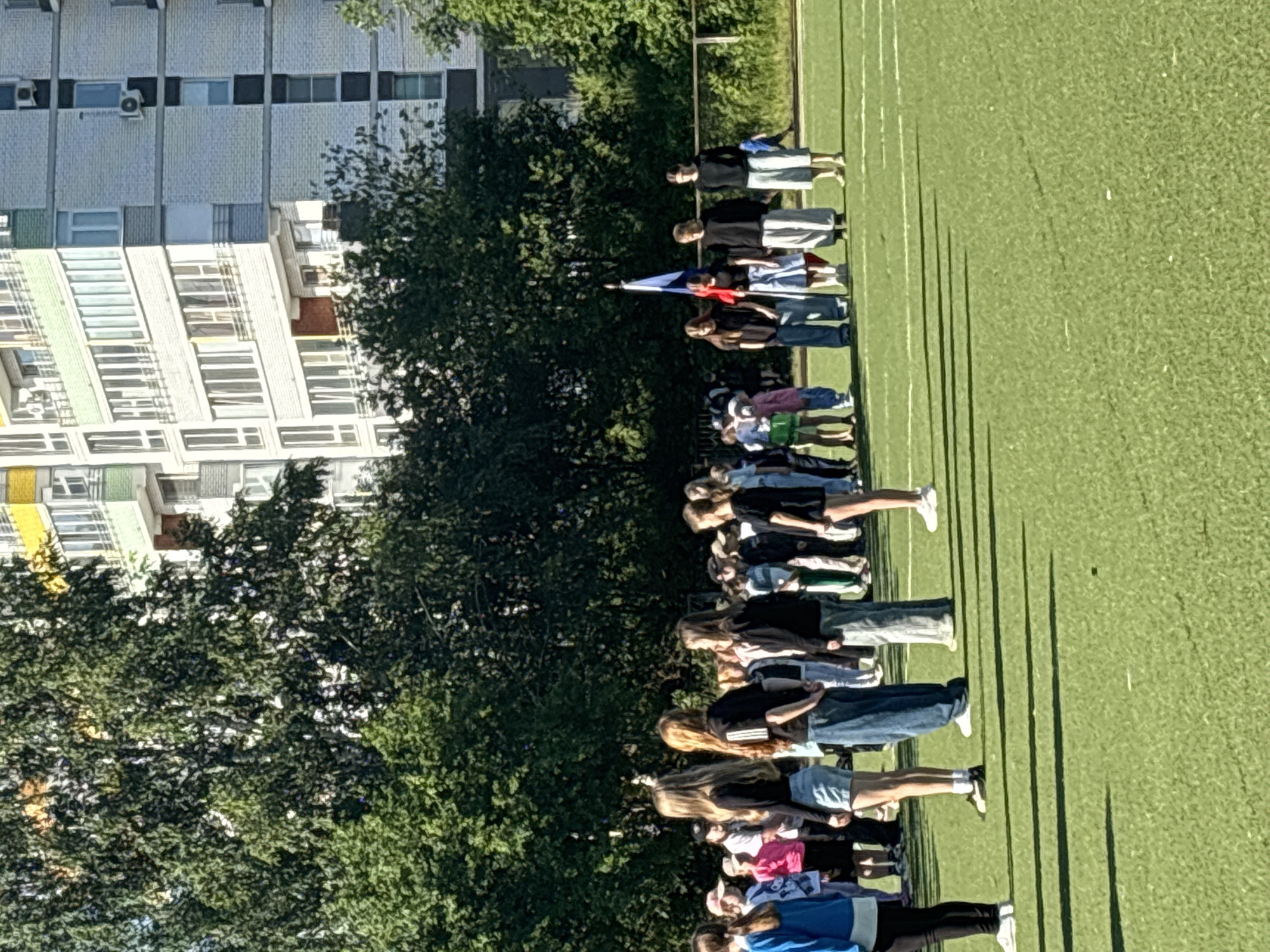 In Chaikovsky, Russia, a group of approximately 30 individuals, appearing to be children and young adults, are gathered on a green artificial turf sports field. They are standing in a line or formation, facing forward. Many are wearing casual clothing such as t-shirts, shorts, and jeans. In the background, a tall, multi-story apartment building with a light-colored brick facade and numerous windows is visible, along with mature green trees. A flagpole with a Russian flag is positioned in the center of the group. The sky is clear and blue, indicating fair weather. The overall scene suggests an outdoor event or gathering, possibly a school or community activity.