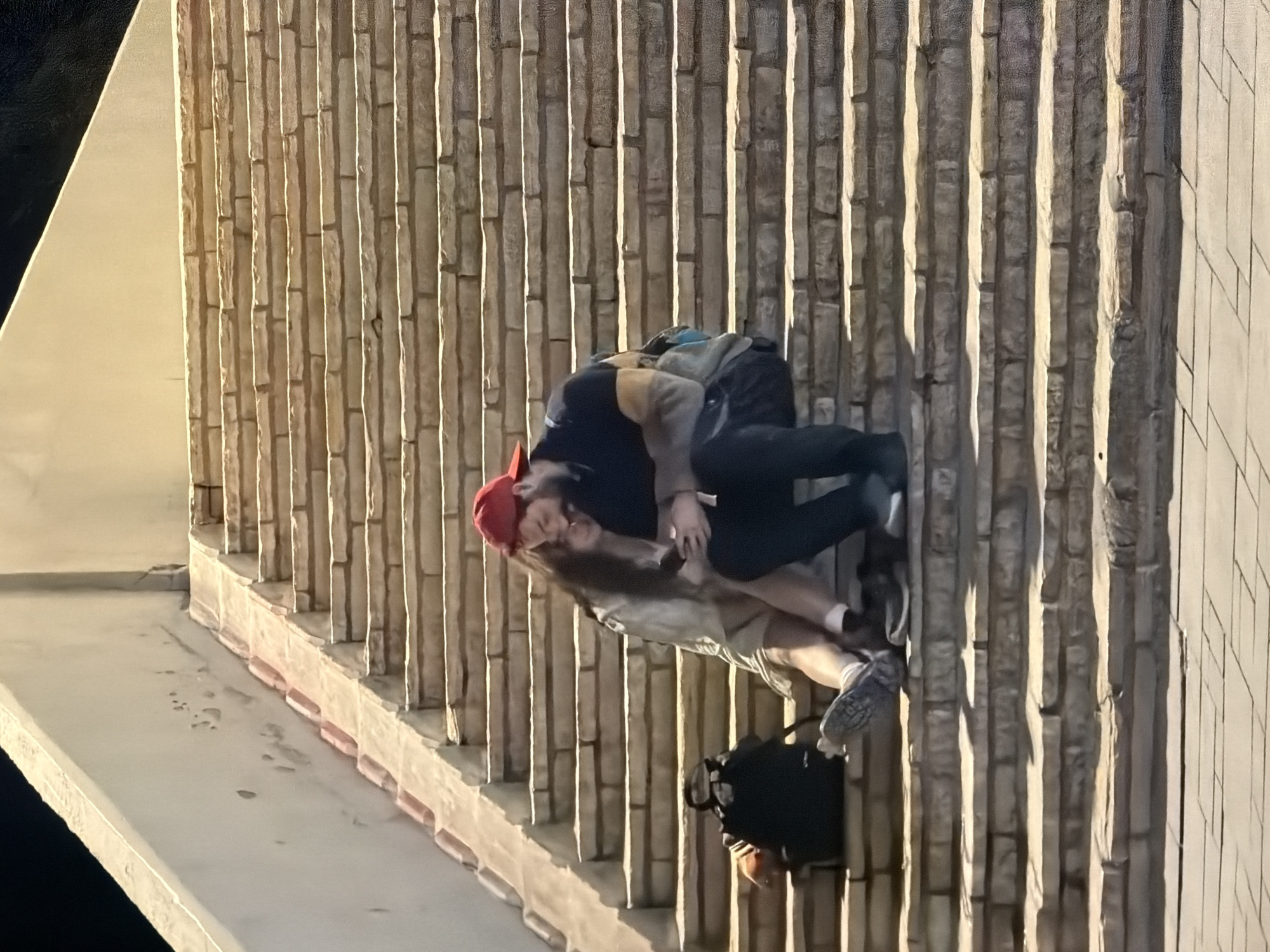 The image shows two individuals embracing beside a textured wall. One person wears a red hat, dark top, and black pants with light-colored shoes. The other person is wearing light-colored shorts and dark shoes. A black backpack is on the ground nearby. The location is identified as Tel-Aviv, Israel, though specific contextual details in the image that identify it as such are not evident. The action depicted is an embrace or hug. The environment is a wall of a building next to what may be a sidewalk.