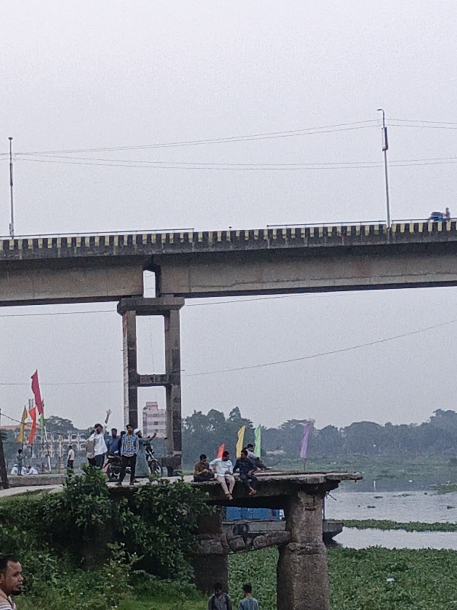 The image captures a scene in Dhaka, Bangladesh, under an overcast sky. A large, modern bridge with a yellow and black striped railing spans the upper portion of the frame. Below the modern bridge is an old, deteriorating stone structure, possibly the remnants of an older bridge or building.
On and around this stone structure, a group of people are gathered. Some are standing, waving, and interacting, while others are sitting on the edge, overlooking a body of water covered with aquatic plants. There are motorcycles parked nearby, suggesting a casual gathering.
In the background, beyond the body of water, there is a building and trees. The foreground features greenery. Some people are walking on a path in front of the greenery. Colorful flags are present, suggesting a festive or commemorative occasion.