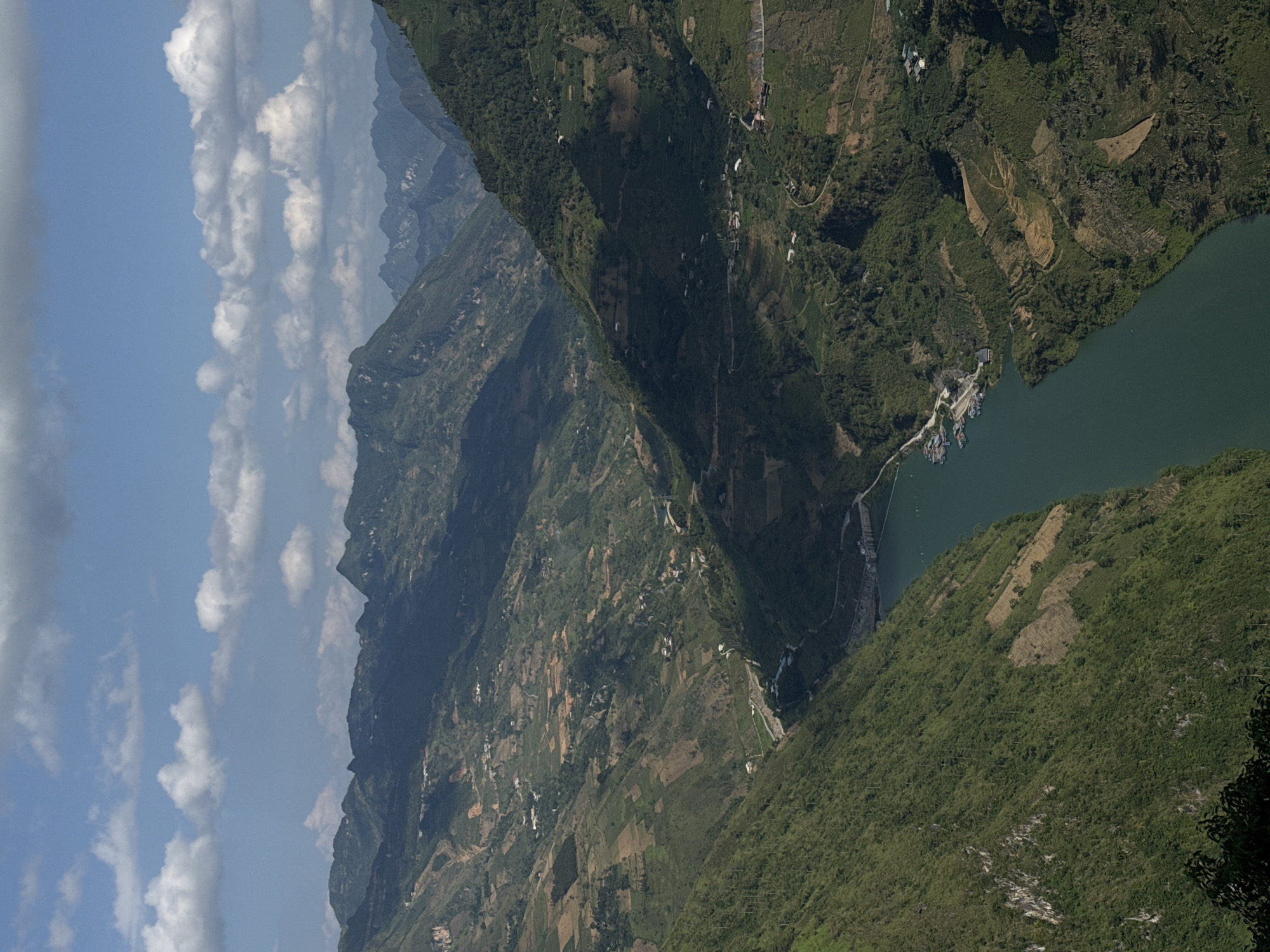 The image shows a mountainous landscape with a river running through it. The location is likely in Đồng Văn Commune, Vietnam. The mountains are covered in lush green vegetation and there are some small villages scattered throughout the area. The sky is blue with white clouds. The river is a dark green color and appears to be calm. There are boats docked in a small bay.