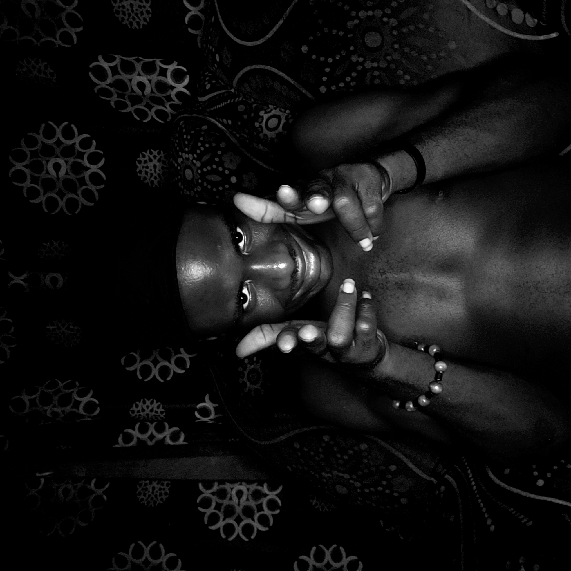 A black and white, high-angle, eye-level shot shows a shirtless person lying on a patterned surface. The person has dark skin and is smiling at the camera, framing their face with their hands. They wear a bracelet on one wrist and a wristband on the other. The surface the person is lying on is patterned with circular and geometric designs. The location, Narayi, Nigeria, is unidentifiable from the image.