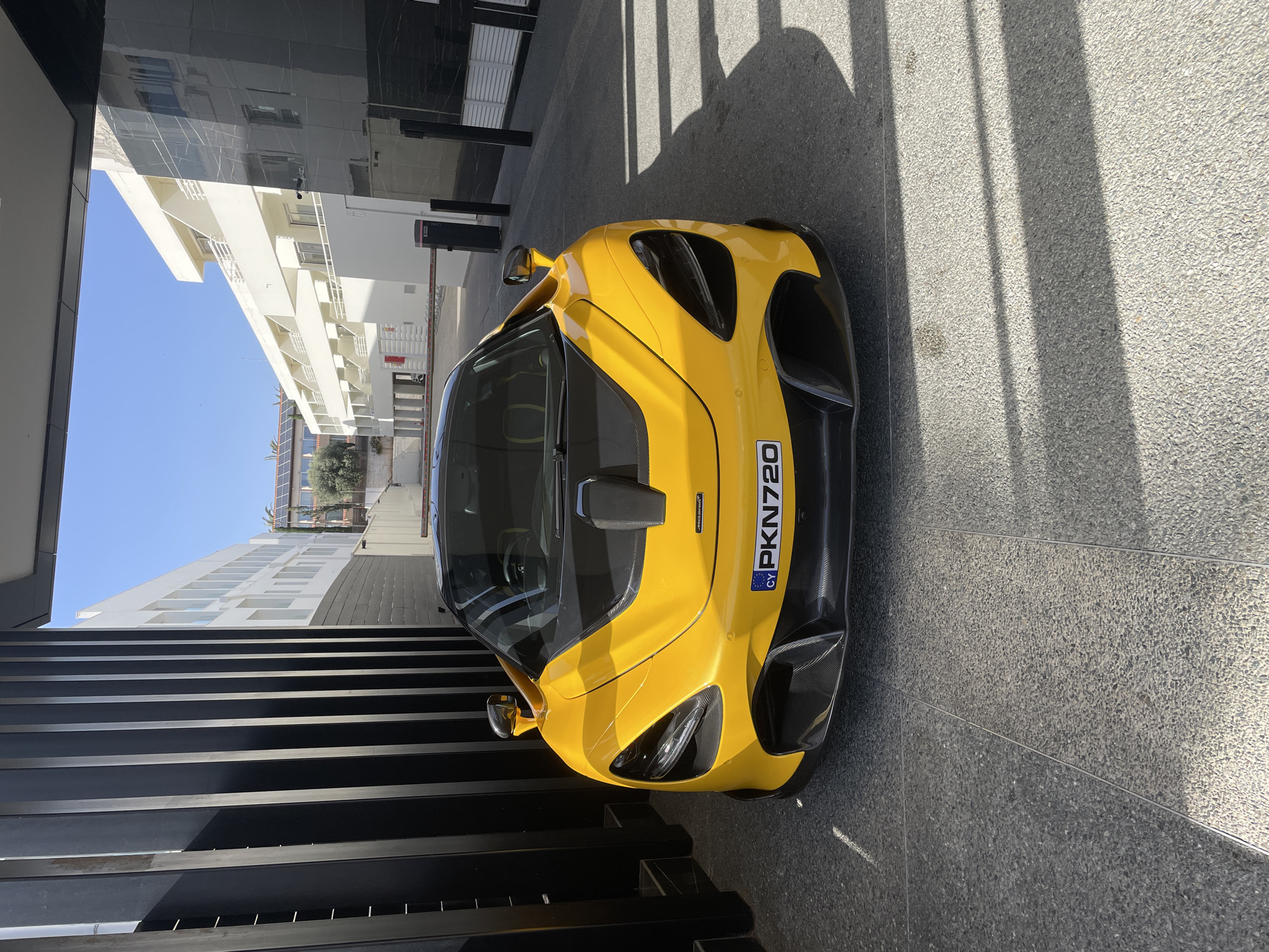 A yellow McLaren sports car with the license plate "PKN720" is parked in front of a modern building in Ayia Napa, Cyprus. The building has a mix of architectural styles, with a dark, mirrored facade and a white section with balconies. Sunlight casts shadows on the ground near the car. There are no visible people in the immediate vicinity.