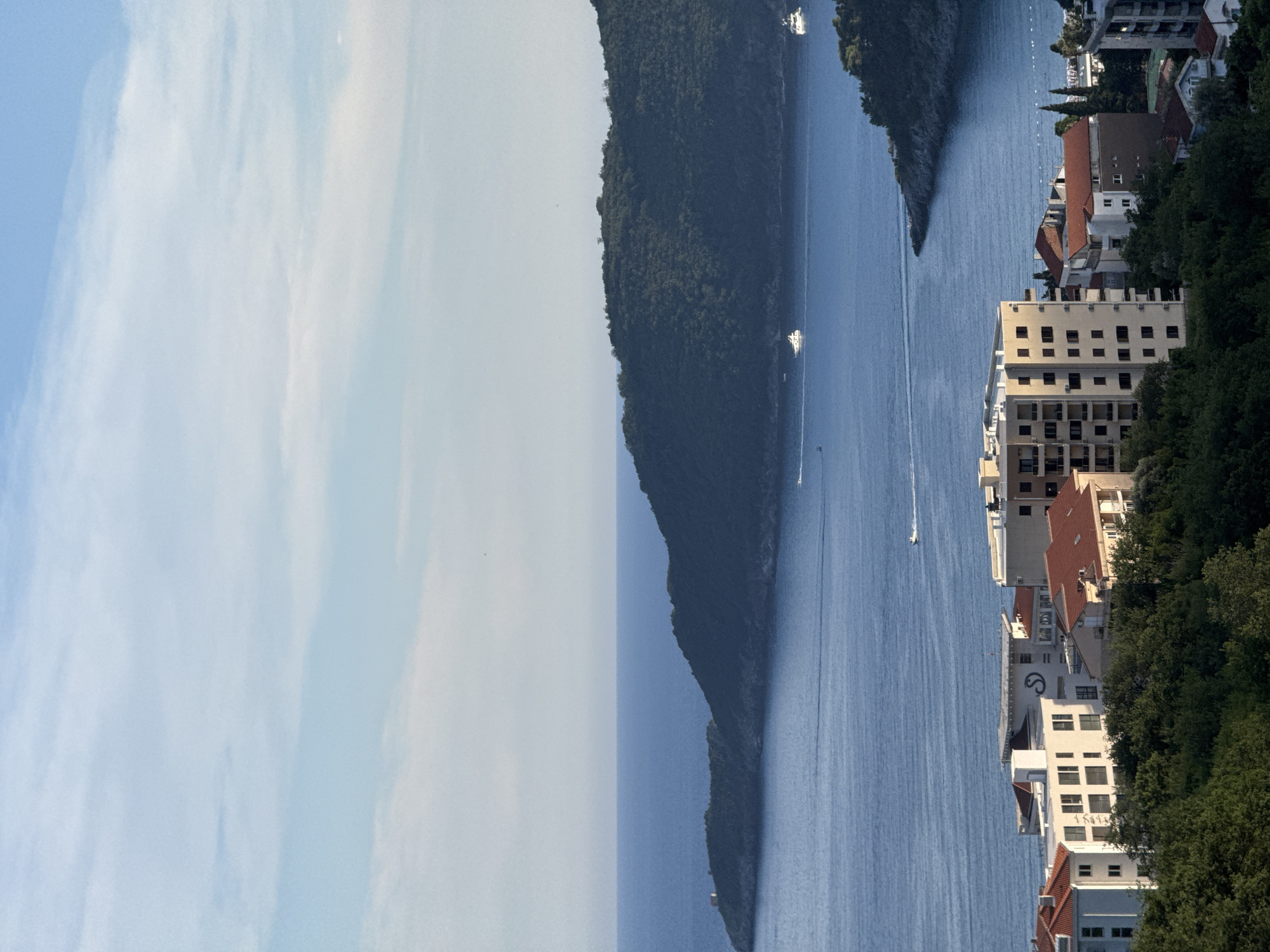 The image captures a coastal scene in Bečići, Montenegro, under a lightly clouded sky. In the foreground, a cluster of buildings with red-tiled roofs and modern apartment blocks are nestled amongst lush green trees. Further back, the calm, blue Adriatic Sea stretches out, with several yachts and smaller boats navigating its waters. The coastline is characterized by a verdant, forested headland that juts into the sea, creating a natural cove. A small structure, possibly a lighthouse or dwelling, is visible on the tip of the farthest peninsula. The overall impression is one of a tranquil Mediterranean landscape, combining urban development with natural beauty.