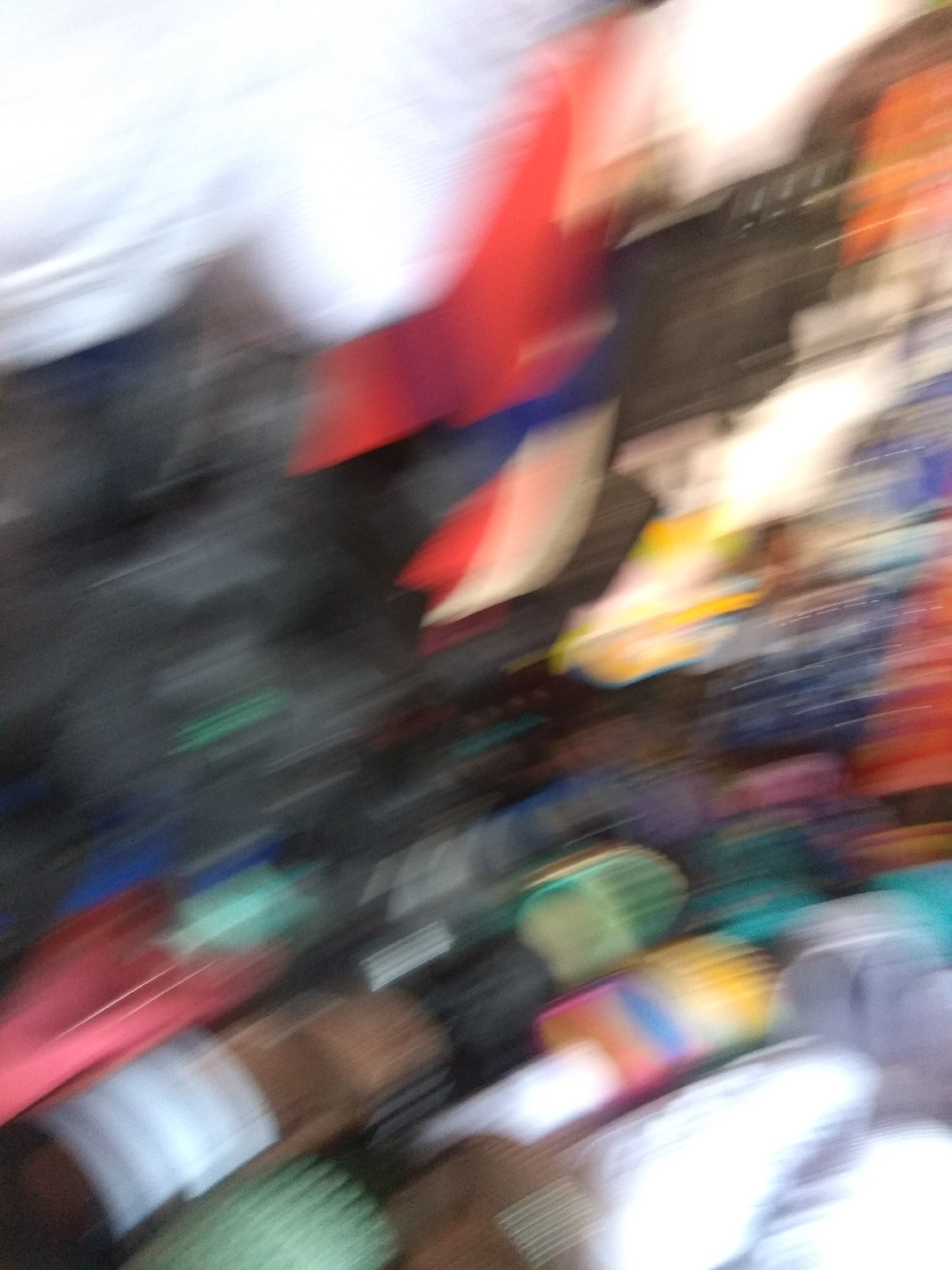 The image is a very blurry, motion-filled snapshot taken in Jalingo, Nigeria. It appears to be a scene with many people and objects, possibly a market or a busy street. The blurriness suggests rapid movement, either of the camera or the subjects.
**Subjects:** The primary subjects are indistinct figures and a variety of objects. There are hints of clothing in various colors, including white, red, blue, and darker shades. Some items appear to be fabric or perhaps merchandise. The overall impression is one of a crowded environment.
**Activity or event:** The motion blur indicates an active scene. It's difficult to pinpoint a specific activity due to the lack of clarity, but the density of subjects and objects suggests a bustling place where people are moving around, perhaps shopping, interacting, or commuting.
**Notable details:** Due to the extreme blur, specific details about time of day, weather, or precise location cues are impossible to discern. No emotions are discernible. The context of Jalingo, Nigeria, provides a general geographic location.
**Visible text:** There is no visible text in the image.