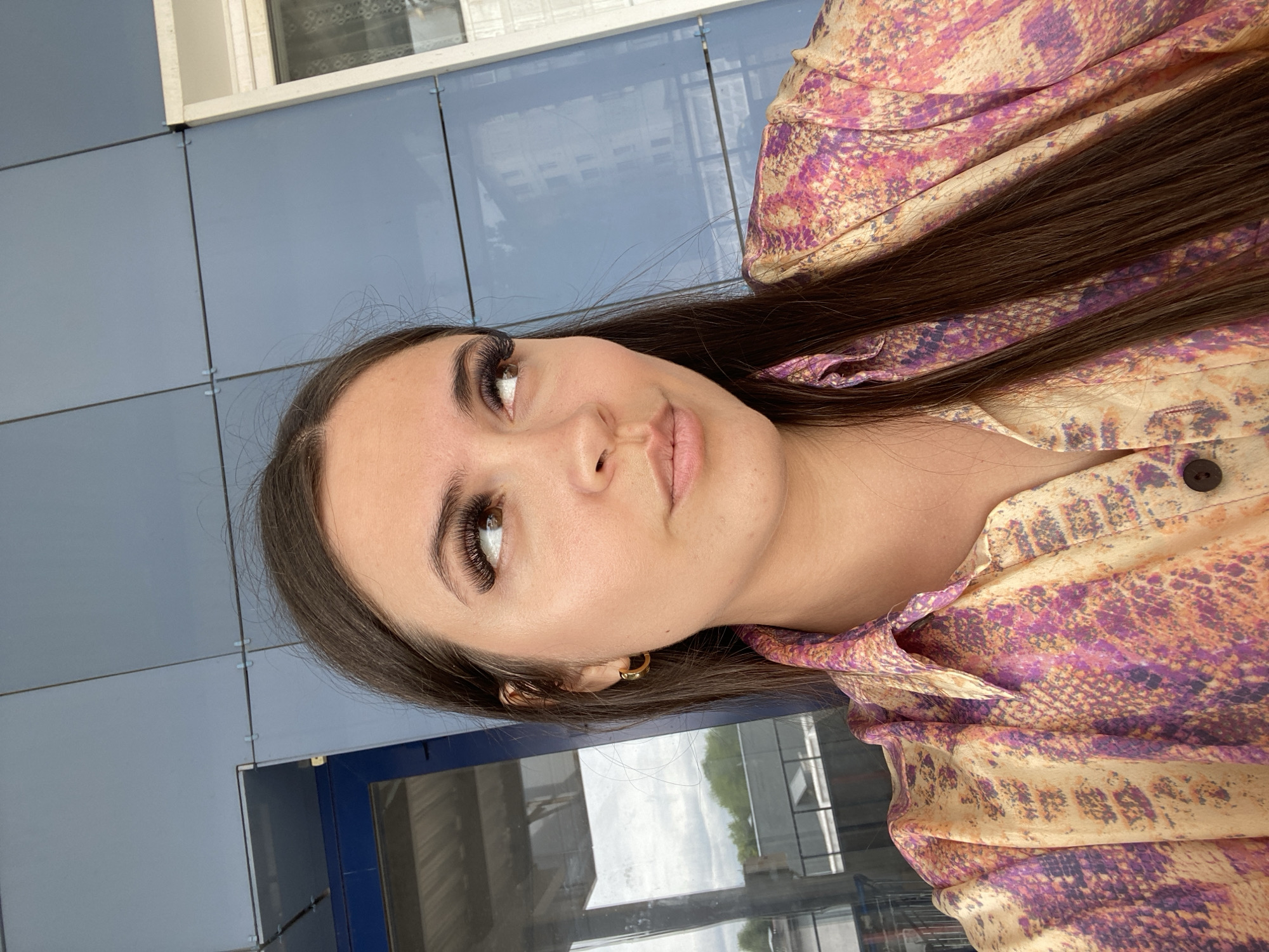 A young woman with long, dark brown hair is positioned in the foreground of the image, facing towards the left. She is wearing a vibrant, collared shirt with a colorful, snake-skin-like pattern in shades of yellow, pink, and purple. A gold hoop earring is visible in her left ear. Her makeup includes dramatic, long eyelashes and subtle lip color.
The background appears to be an urban exterior. The prominent feature is a wall made of large, rectangular blue panels, likely part of a modern building. To the right of the blue wall, a white frame surrounds a window, through which a building is partially visible. To the left of the blue wall, a large glass pane reflects the sky and some trees, suggesting the presence of a building with glass facades or windows. The overall setting hints at a street or public space in the city of Chaikovsky, Russia, characterized by modern architecture and possibly a commercial or residential area. The lighting suggests it is daytime, with natural light illuminating the scene.