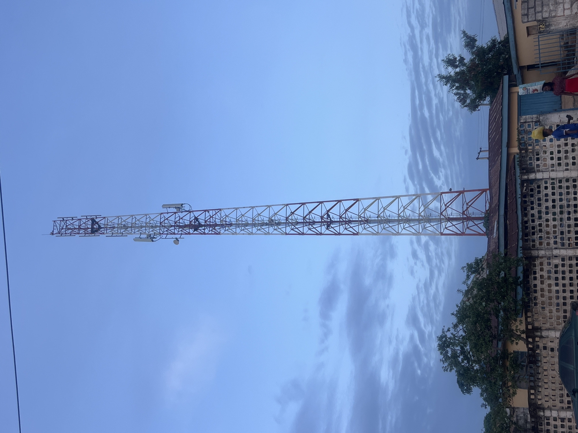 A tall, lattice-style telecommunications tower dominates the central-left of the image, extending vertically against a pale blue sky. The tower is constructed from metalwork, painted in alternating sections of reddish-brown and white or light grey, and is equipped with various antennas and dishes along its structure.
On the right side of the image, the upper parts of residential or commercial buildings are visible. These buildings feature concrete block walls, some with decorative patterned ventilation blocks, and corrugated metal roofs. A green metal gate is partially seen between two structures. In the foreground, a vibrant green tree with dense foliage grows near the buildings.
Several people are discernible near the buildings and gate. One individual is wearing a red top and another has a yellow head covering, while other figures are less distinct. They appear to be standing or moving casually.
The sky is partly cloudy, displaying elongated, streaky cloud formations that suggest either early morning or late afternoon light. The overall atmosphere is calm and clear. A faint, thin cable or power line runs vertically along the far left edge of the frame. The number "73" is clearly visible on the wall of one of the buildings, near the green gate. The scene depicts an urban or semi-urban environment in Jos, Nigeria, characterized by local architecture and infrastructure. No specific event or major activity is unfolding.