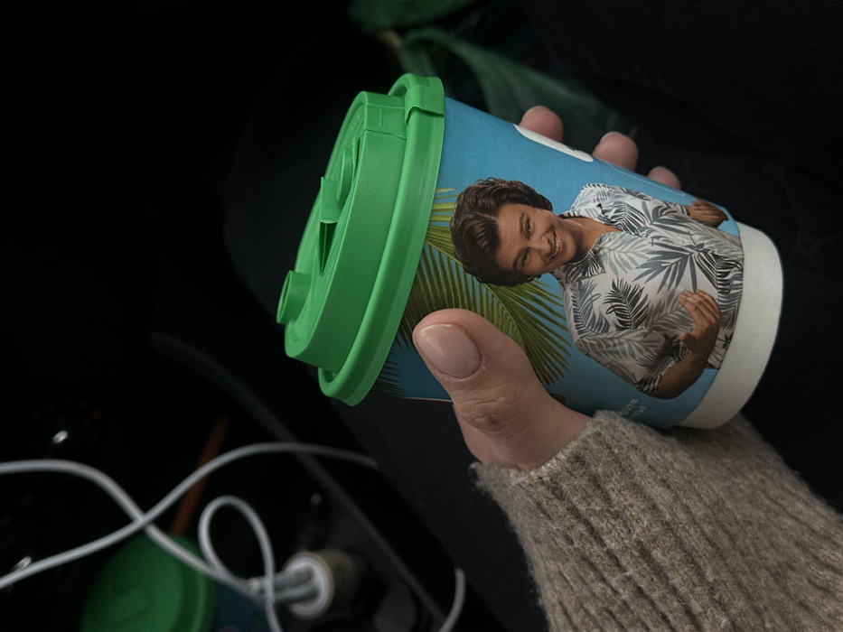 The image depicts a right human hand holding a disposable beverage cup inside the interior of a vehicle, located in Starkovo, Russia. The hand, with pale skin and manicured nails painted in a light nude shade, is partially covered by a light brown knitted sweater sleeve. The paper cup is light blue, decorated with green palm frond illustrations, and features a smiling man with wavy brown hair wearing a patterned short-sleeve shirt. The cup is sealed with a bright green plastic lid that includes a drinking flap. In the out-of-focus upper left background, a white charging cable connects to a power adapter plugged into what appears to be a car's center console, next to a partially visible second beverage cup with a green lid.