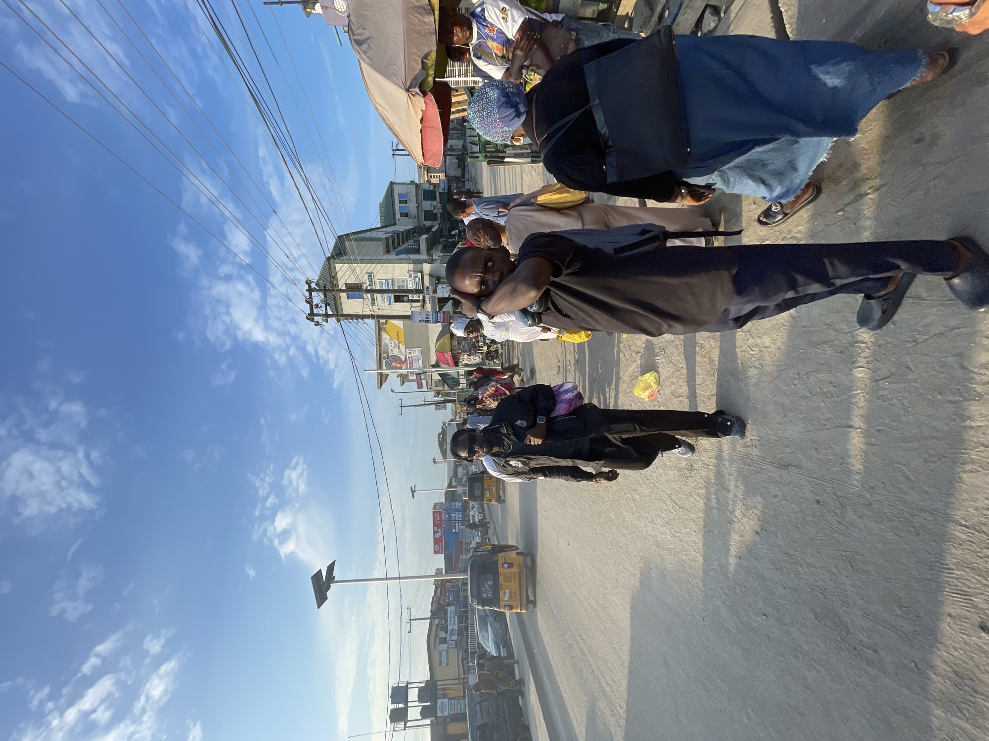 The image depicts a bustling street scene in Lagos, Nigeria, likely during the late afternoon given the long shadows cast across the pavement. The sky is a clear blue with scattered white clouds. The scene is oriented vertically, with the ground appearing at the top of the image.
Several people are visible, some walking and others gathered near market stalls. In the foreground, a person wearing a black t-shirt and dark pants is looking towards the camera, with headphones around their neck. Further back, a group of people are seated under a canopy, possibly at a market stall or a waiting area. One individual wears a blue cap with a patterned design. Another person in the mid-ground is dressed in a dark jacket and light-colored trousers, with a backpack slung over their shoulder. In the background, several vehicles, including a yellow auto-rickshaw, are on the road, suggesting moderate traffic. Buildings line the street, with signage indicating businesses and shops.
The overall atmosphere appears lively and dynamic, typical of a busy urban environment. The sunlight indicates it is daytime. The presence of auto-rickshaws and the architectural style of the buildings are strong cues for the location being Lagos, Nigeria. Visible text includes signs on buildings and possibly advertisements on vehicles, though the text is largely illegible due to resolution and orientation.