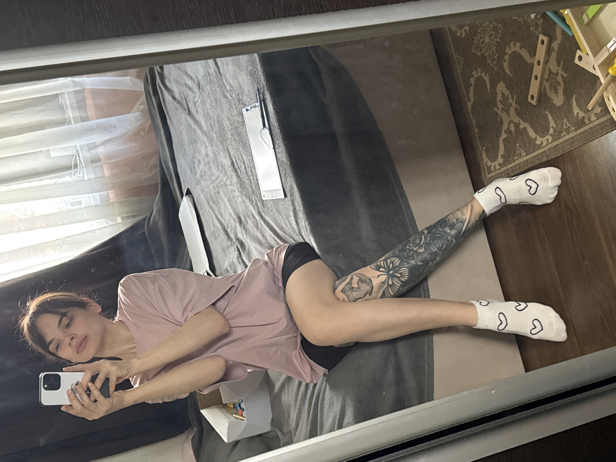 A woman with brown hair is visible in a mirror selfie. She is lying on a gray bed with a white box and a silver object on it. She is wearing a pink t-shirt, black shorts, and white socks with black hearts on them. She has tattoos on her left leg. A window with white blinds is to the left of the bed. A rug and children's toys are on the floor to the right of the bed. The city of Rostov-on-Don, Russia is not discernible in the image.